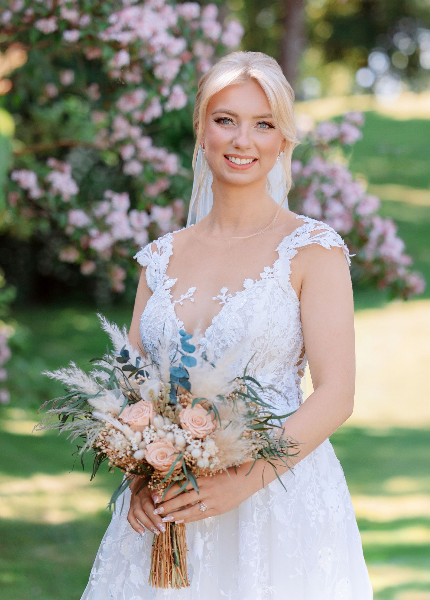 A bride in a white lace gown holds a delicate bouquet of peach and blush flowers with eucalyptus in a garden setting.