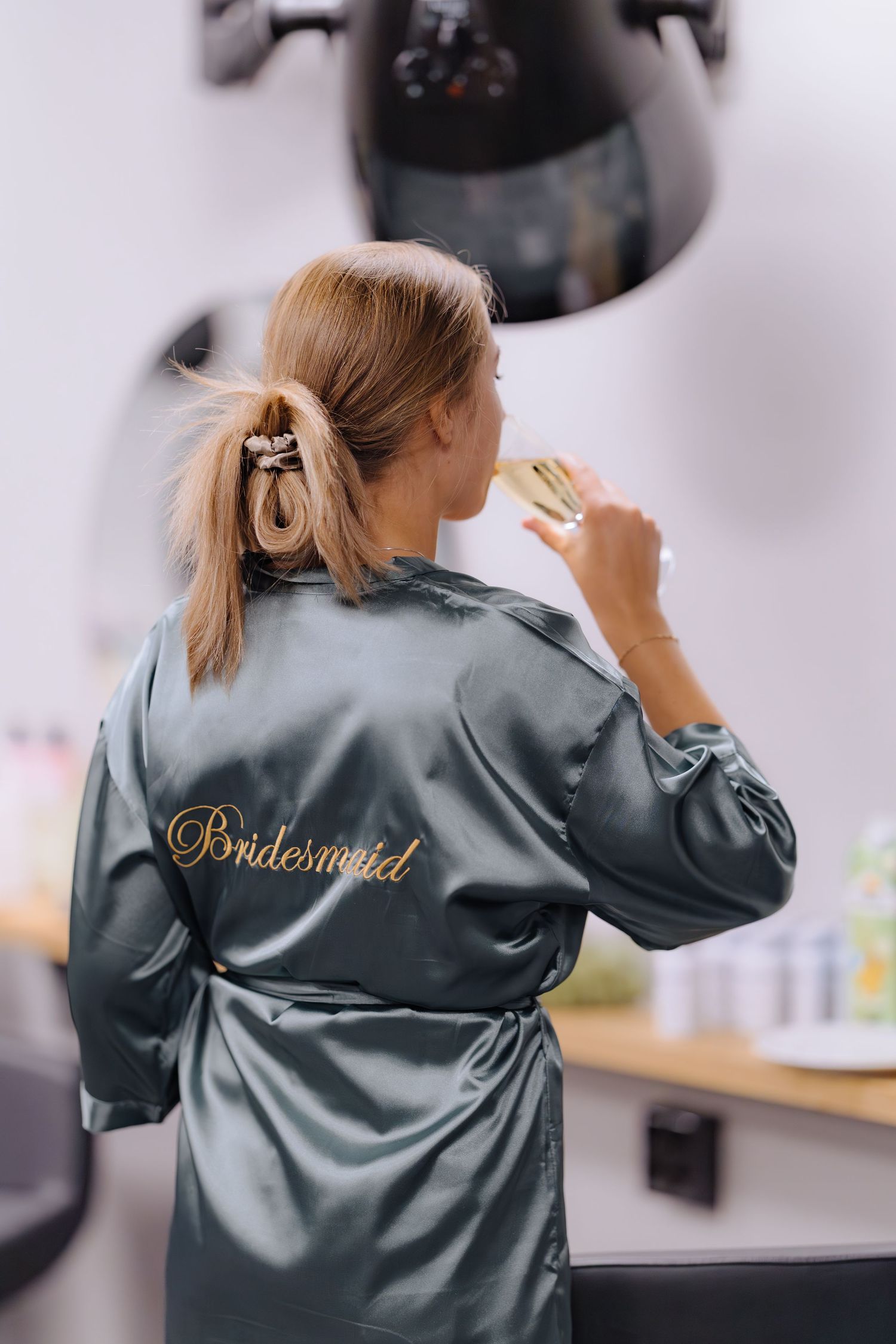 A bridesmaid in a silky grey robe sits at a vanity table getting ready for a wedding celebration.
