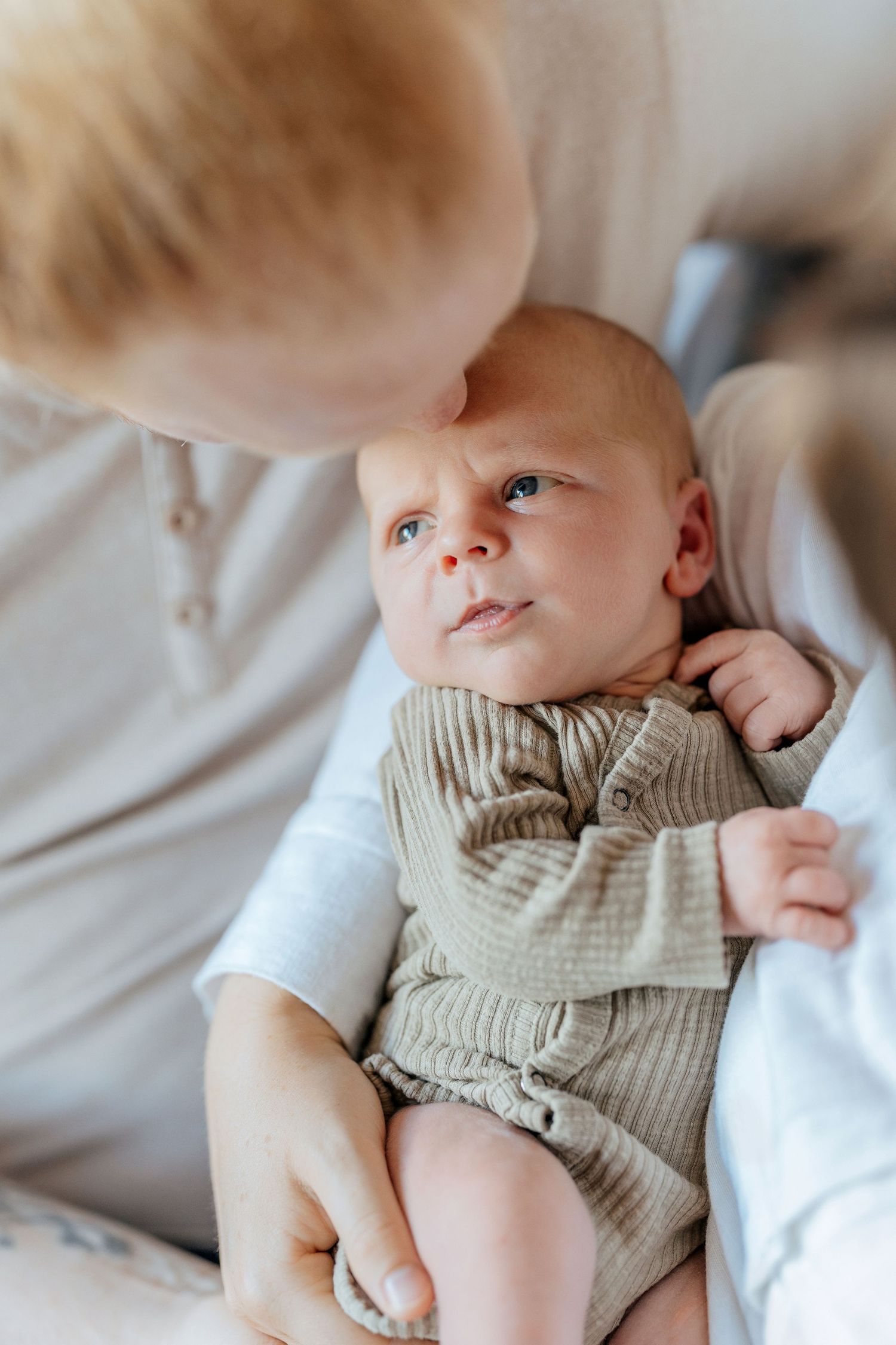 A newborn baby in a sage green ribbed romper gazes upward while being cradled by a blonde person.