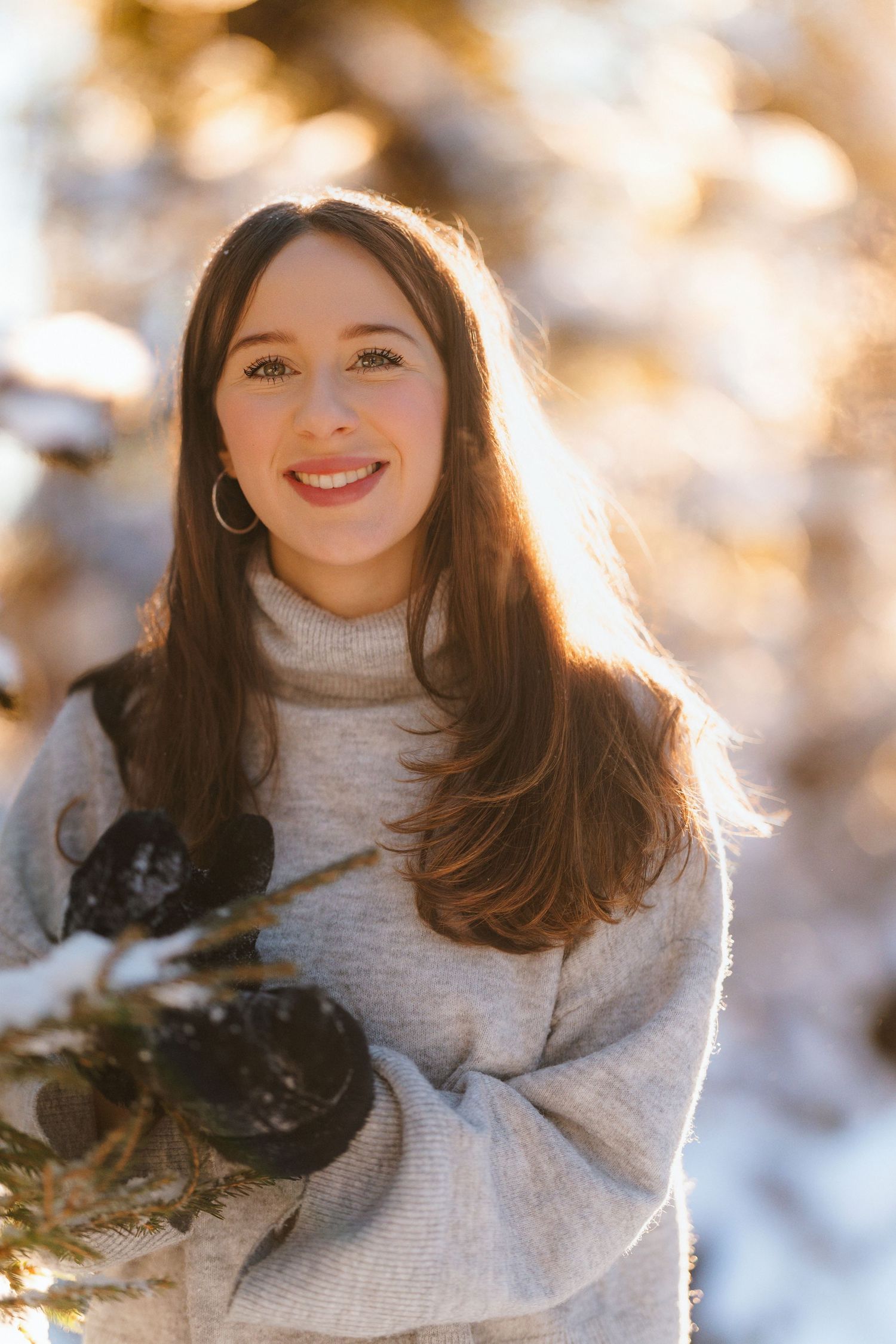 Young woman in gray sweater and black gloves smiling outdoors in snowy winter setting with golden bokeh background.