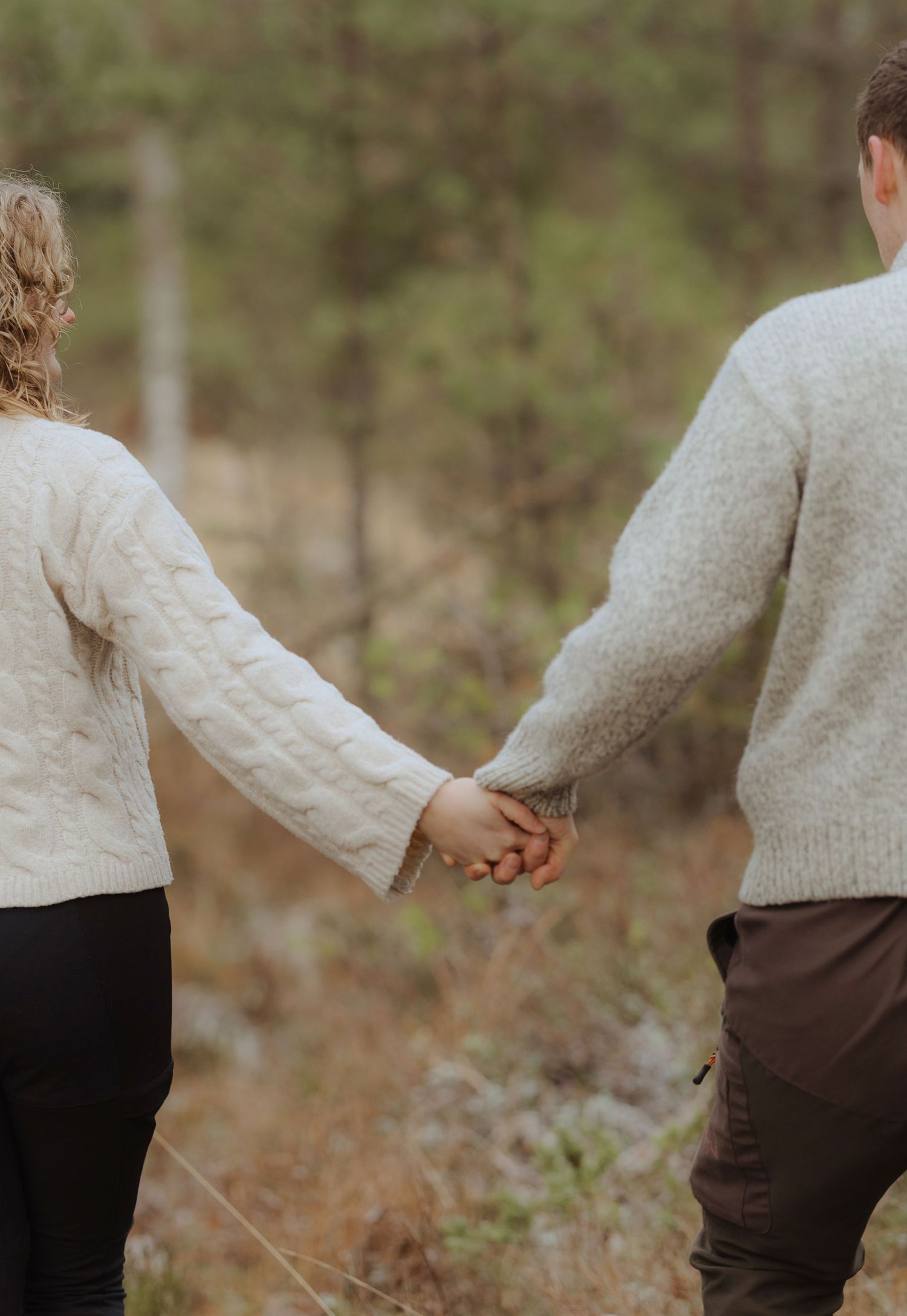 A couple wearing matching cream-colored sweaters hold hands while walking together in a forest setting.