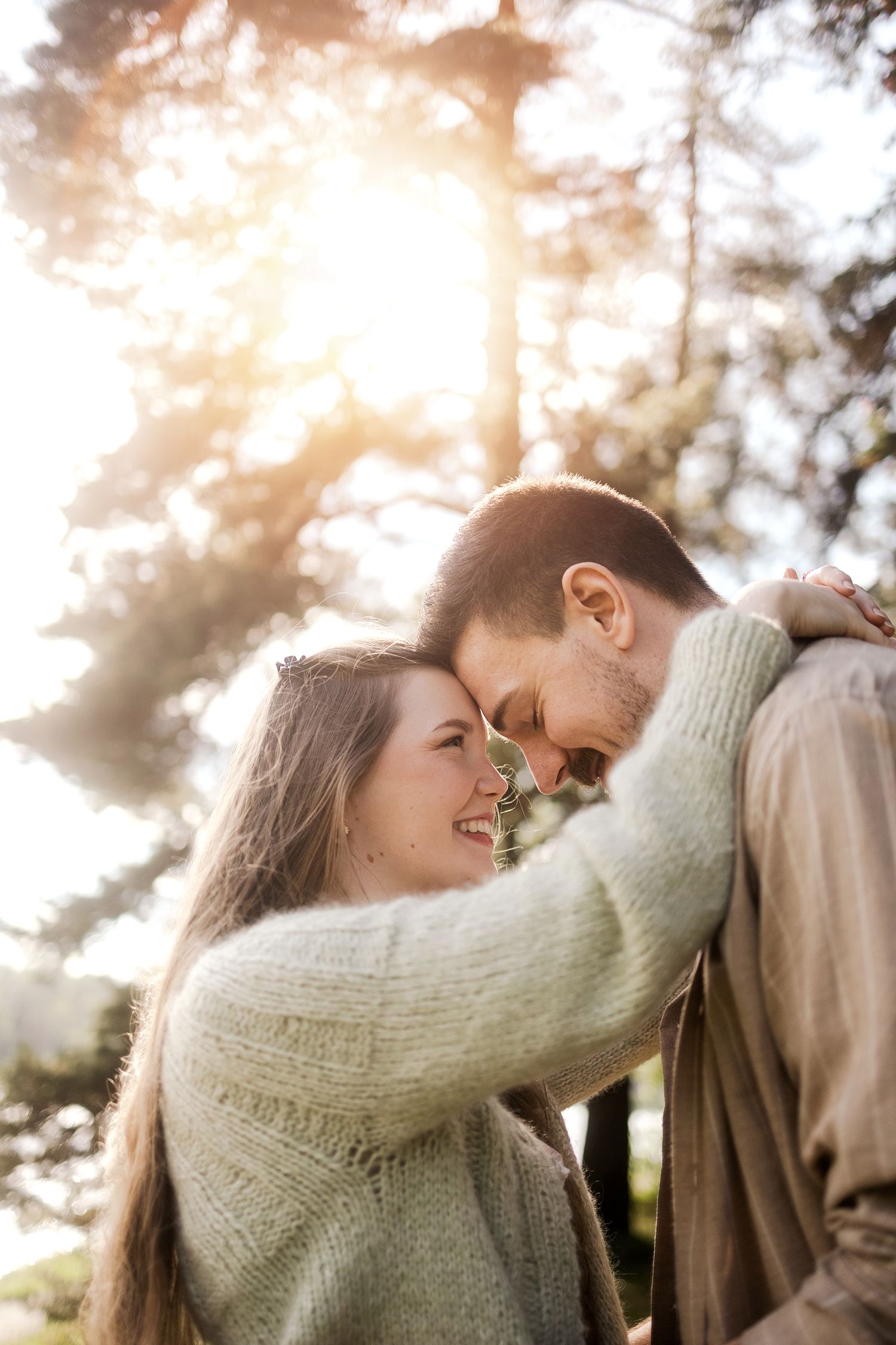 A romantic couple embraces in soft sunlight with evergreen trees in the background.