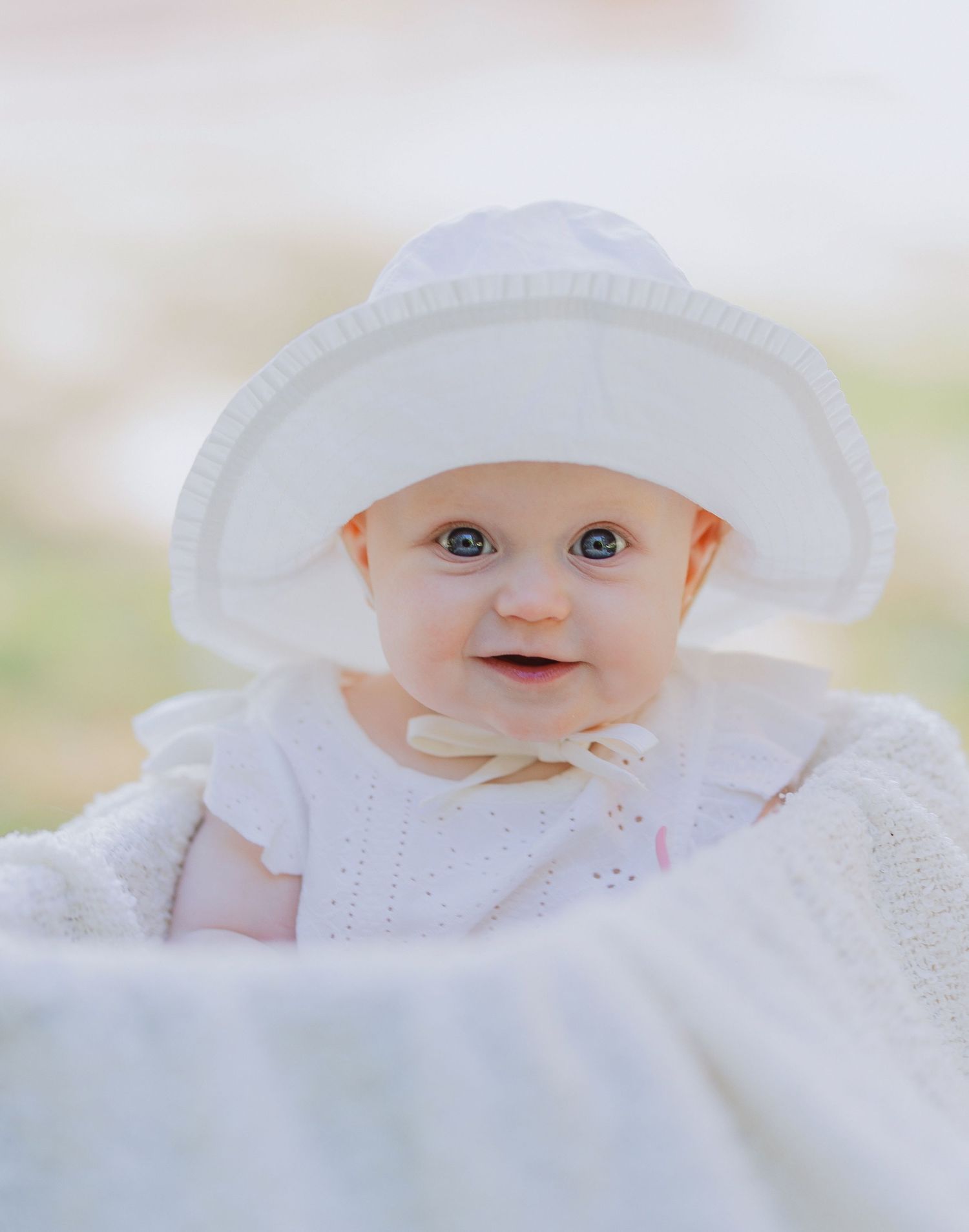 A baby wearing a white sunhat and dress smiles while lying on soft white fabric.