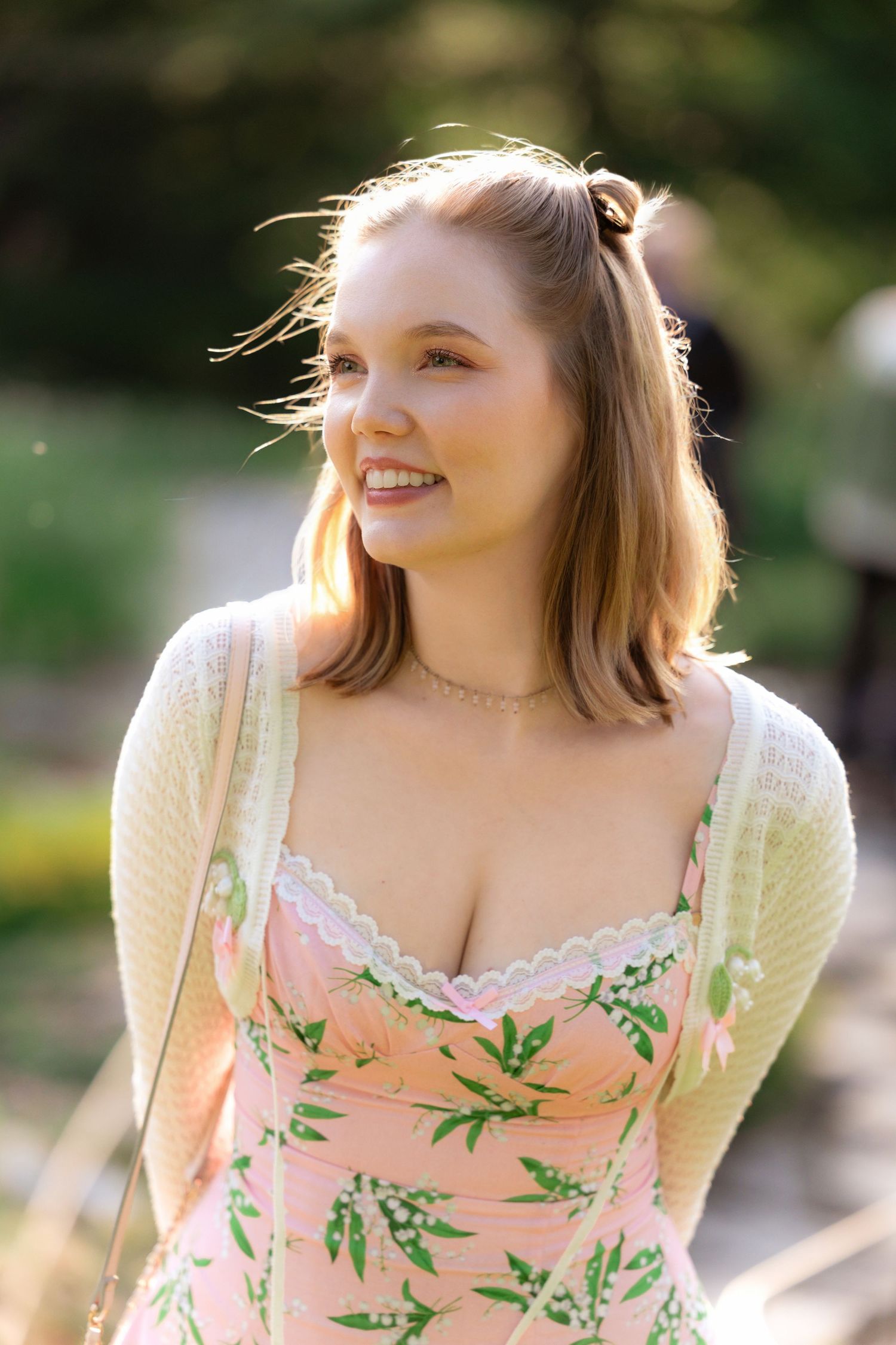 A person wearing a floral peach-colored sundress with a white cardigan poses outdoors in natural sunlight.