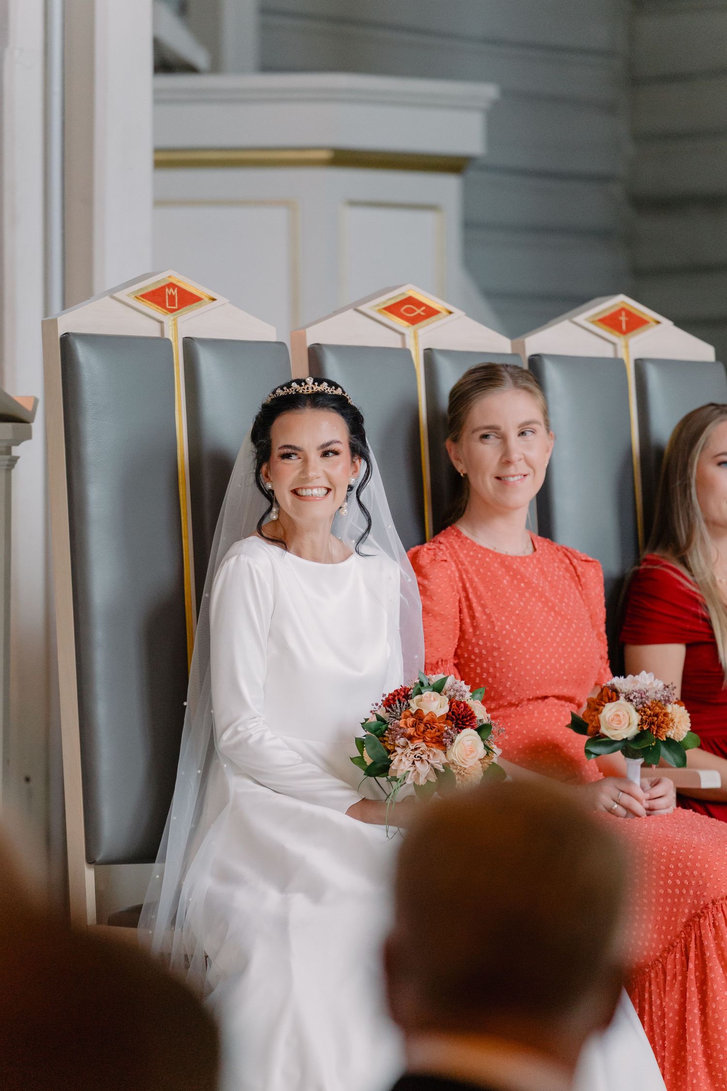 Wedding party members in white and coral dresses stand together in a bright indoor venue during a celebration.