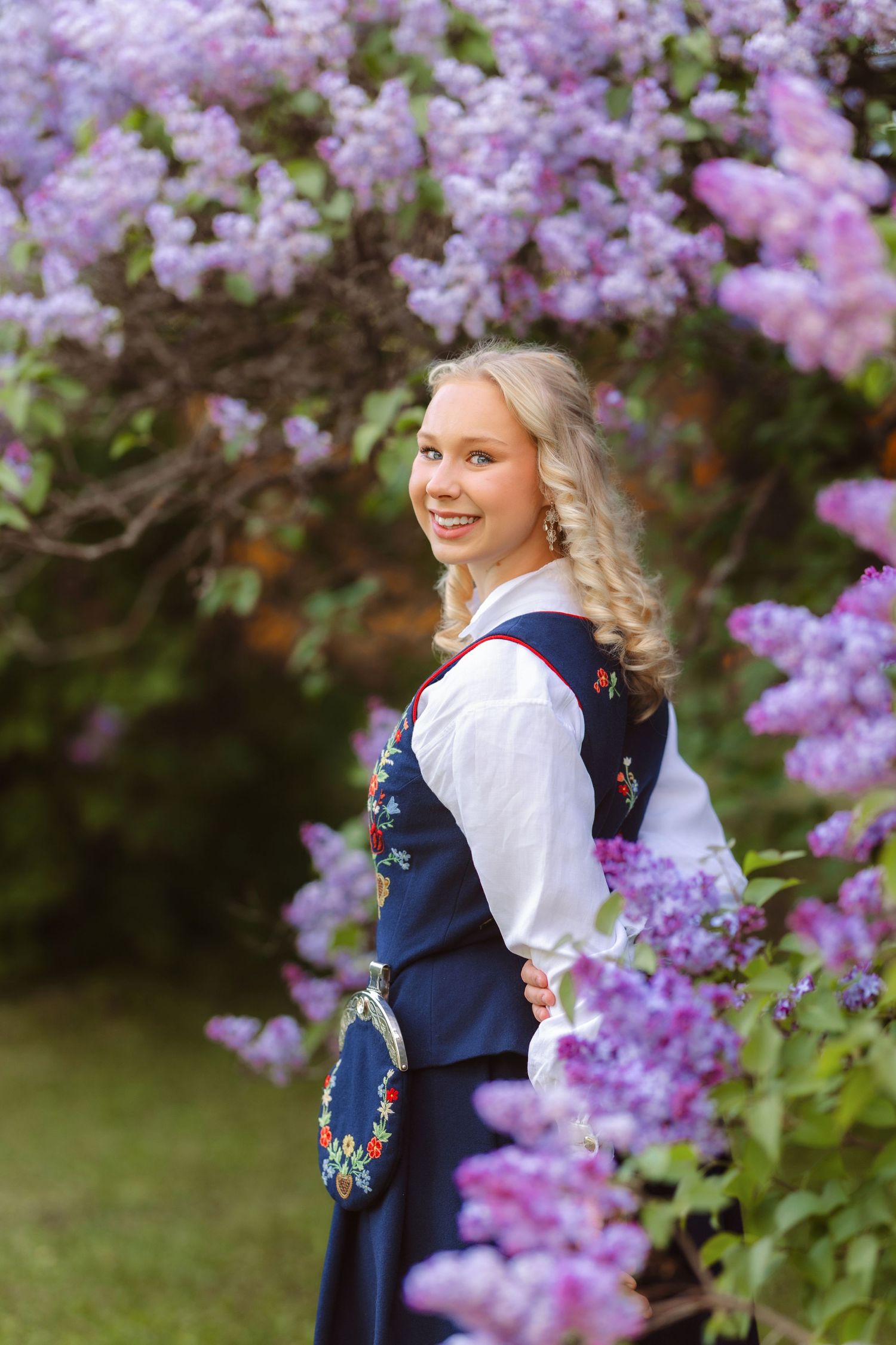 Young blonde woman in traditional Norwegian bunad dress smiling among purple lilac blooms in garden.