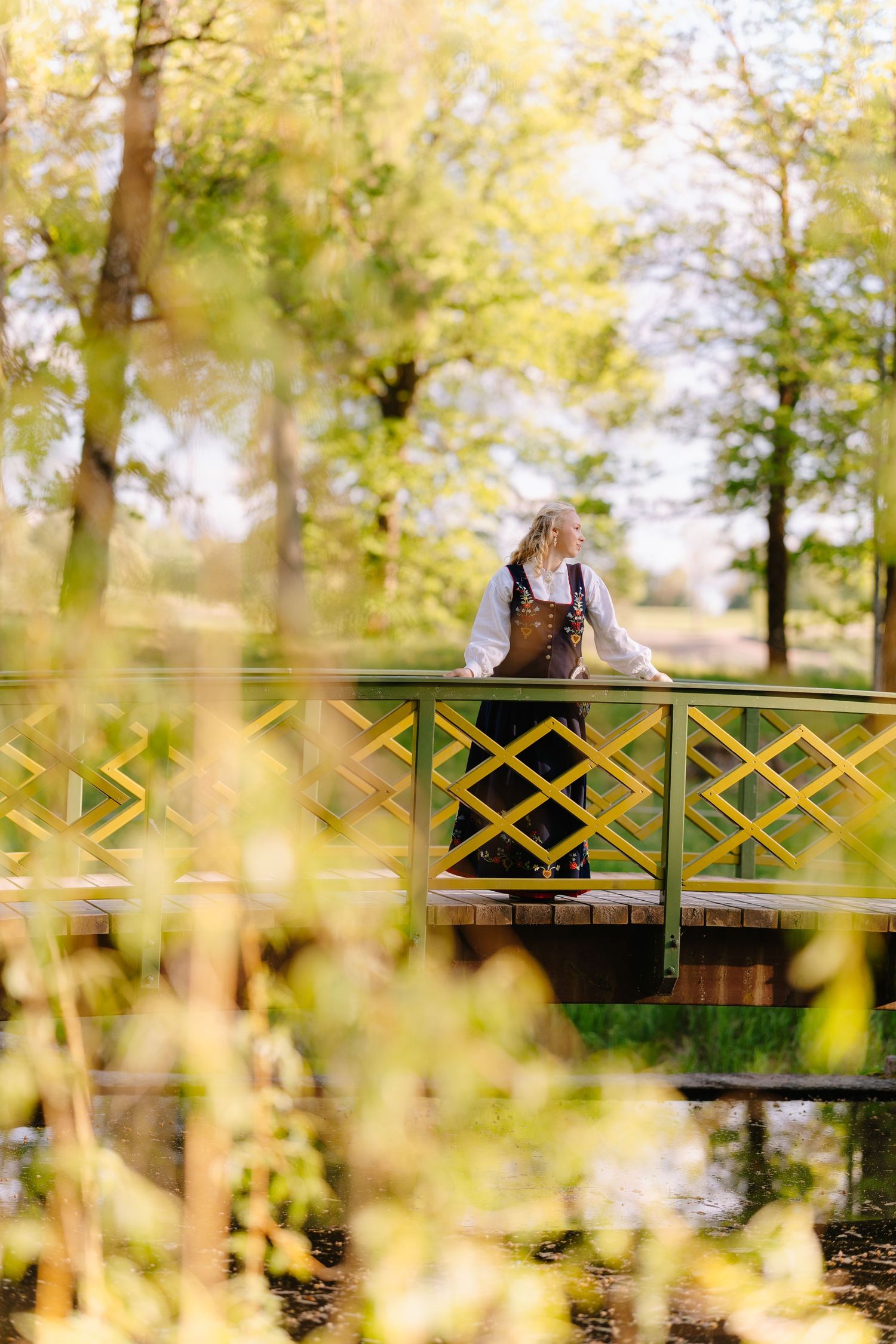 Woman in traditional attire stands on a decorative green bridge surrounded by lush spring trees in a scenic park.