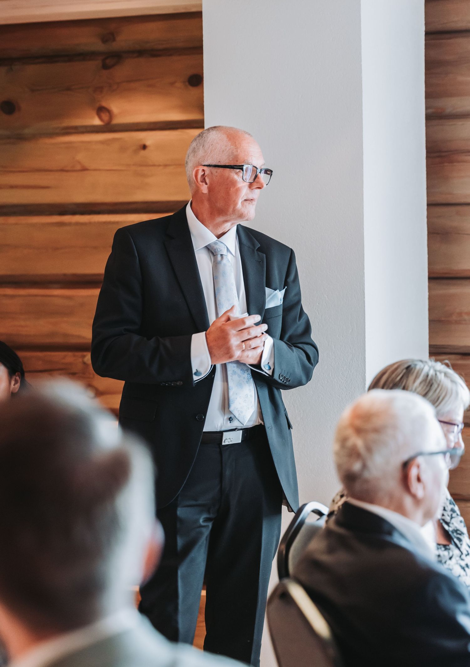 A speaker in a dark suit and tie addresses an audience in a wood-paneled room during a business presentation.