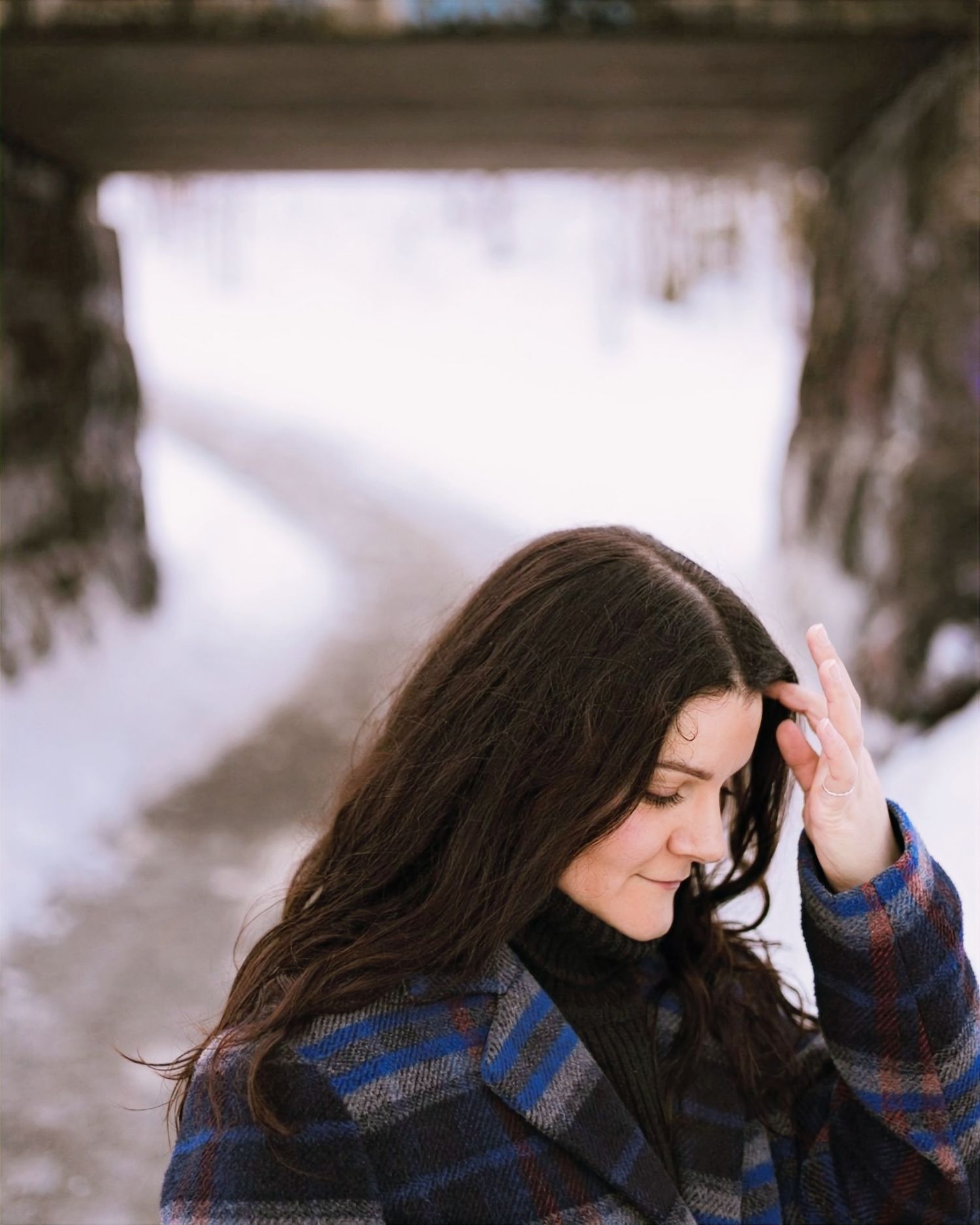Person in a plaid winter coat stands thoughtfully in a snowy outdoor setting with soft lighting.