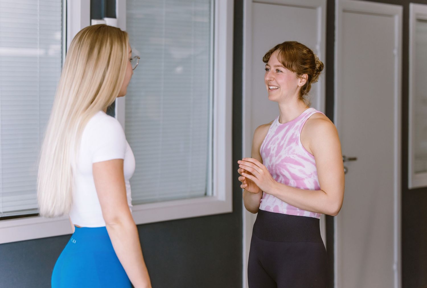 Two women in casual attire stand and chat in a modern office hallway with framed artwork on the wall.