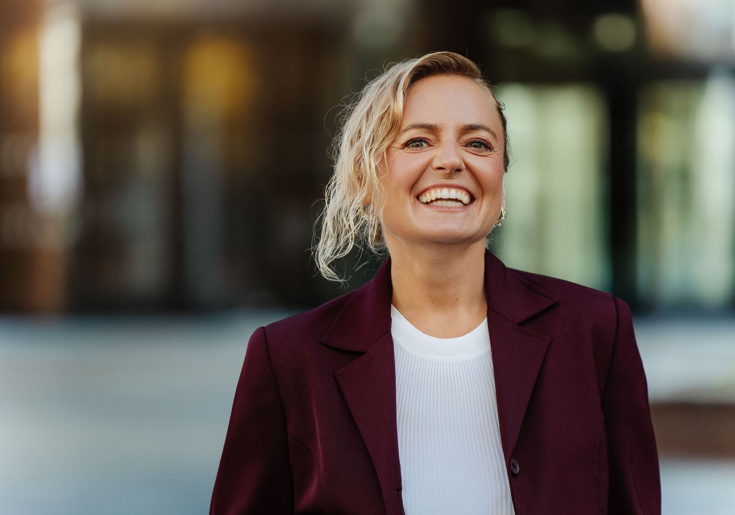 A person in a burgundy blazer and white top smiles while posing against a blurred background.