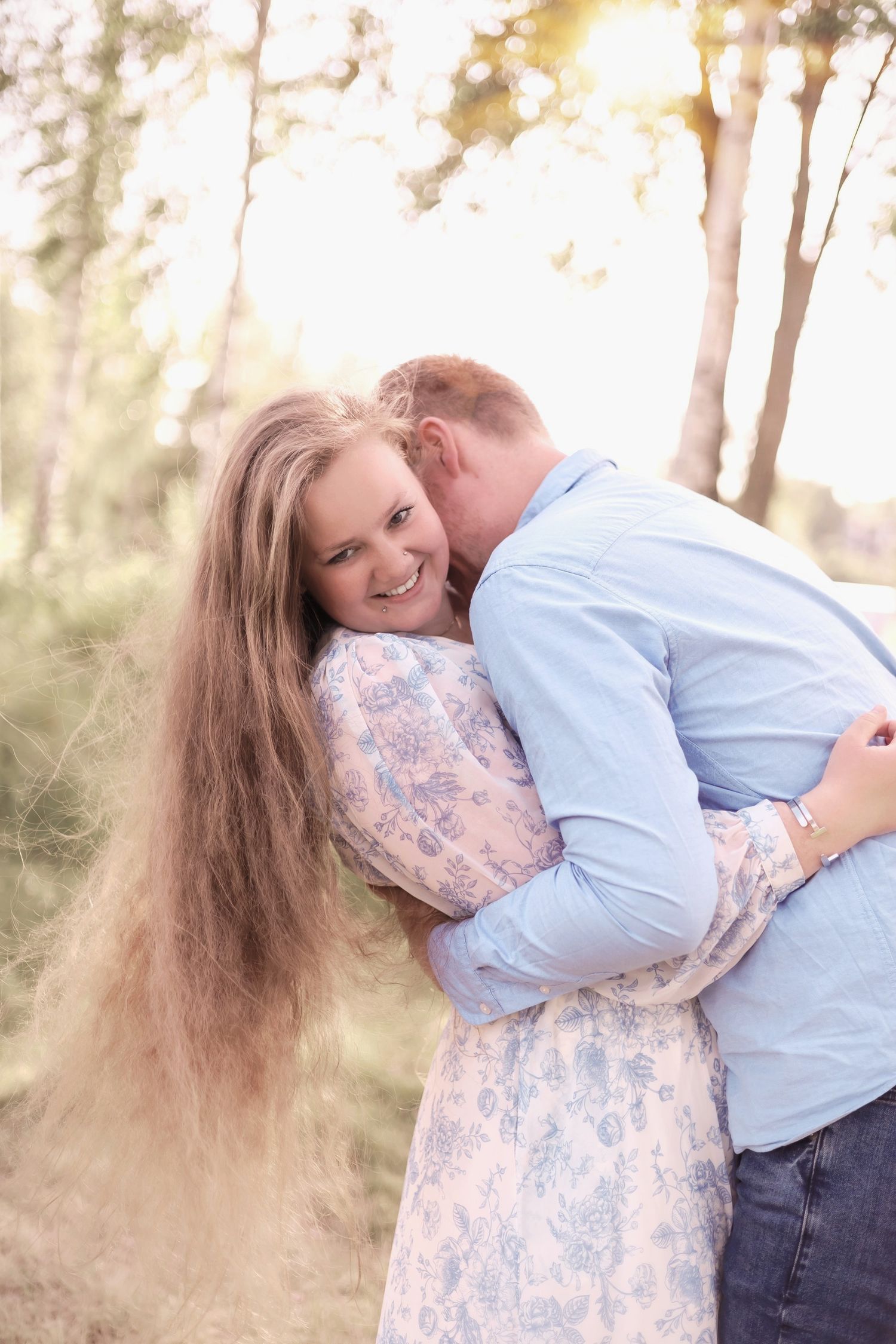 A romantic outdoor couple portrait shows an intimate embrace among trees at sunset with soft golden lighting.