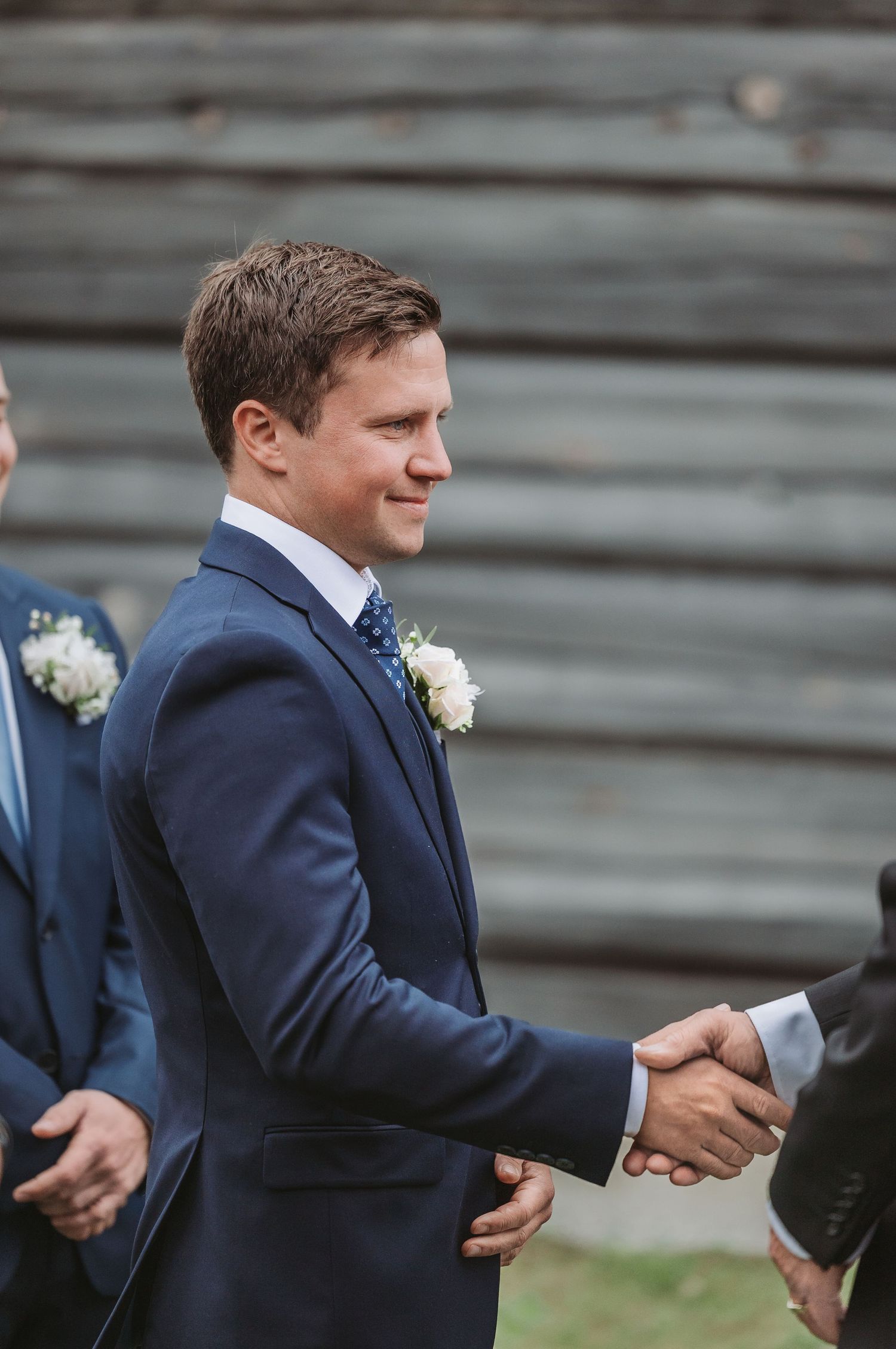 Groomsman in navy blue suit shakes hands with another guest at outdoor wedding ceremony.