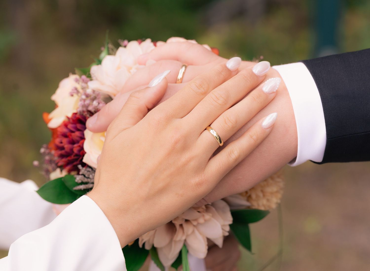 Newlywed couple's hands with wedding rings intertwined over pink and burgundy flowers in romantic pose.
