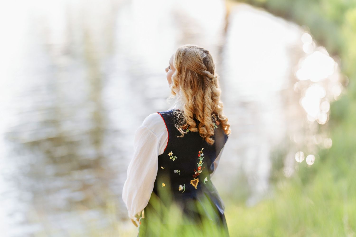 Young woman in traditional embroidered dirndl vest stands by a sunlit lake, her curly blonde hair cascading down.