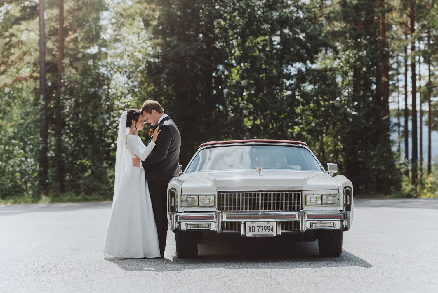 A classic white Cadillac parked in a wooded area serves as a backdrop for a romantic wedding photo session.