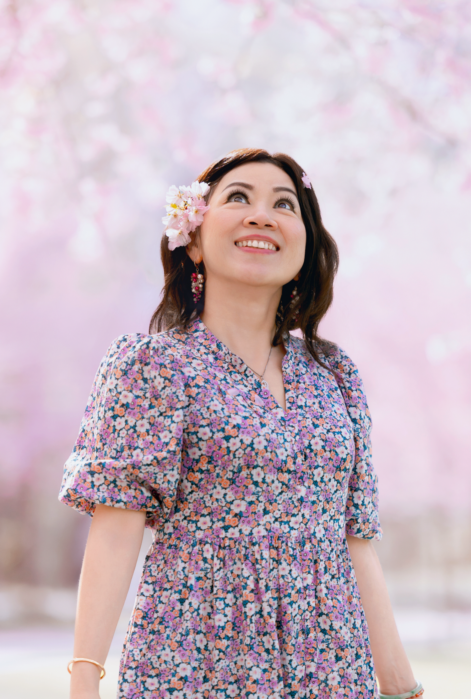 A person in a floral print dress smiles joyfully under blooming cherry blossom trees on a spring day.