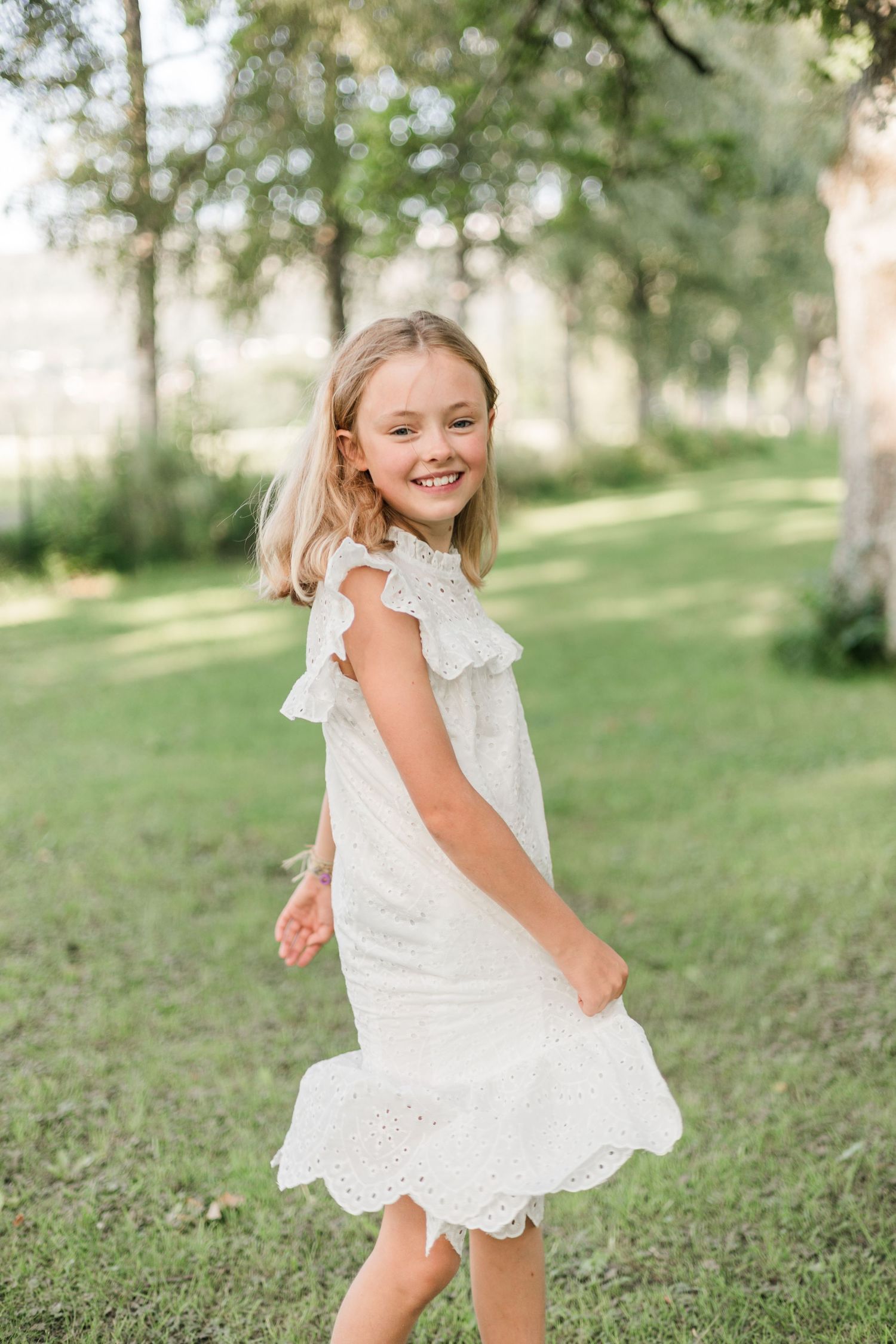A young person in a white eyelet lace dress twirls playfully on a grassy hill with trees in the background.