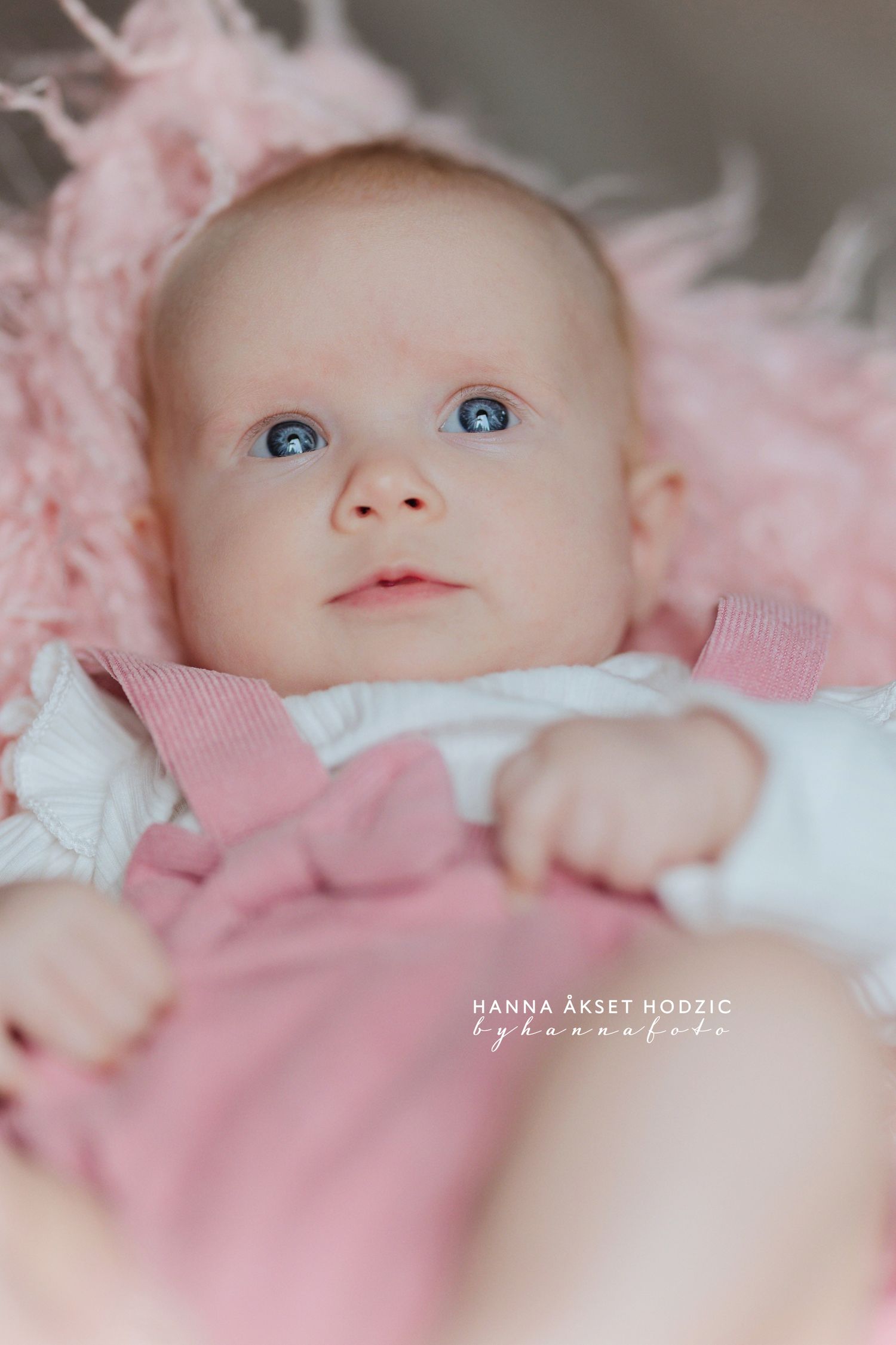 A baby with bright blue eyes wearing pink and white clothing lies on soft pink fabric in a professional portrait.