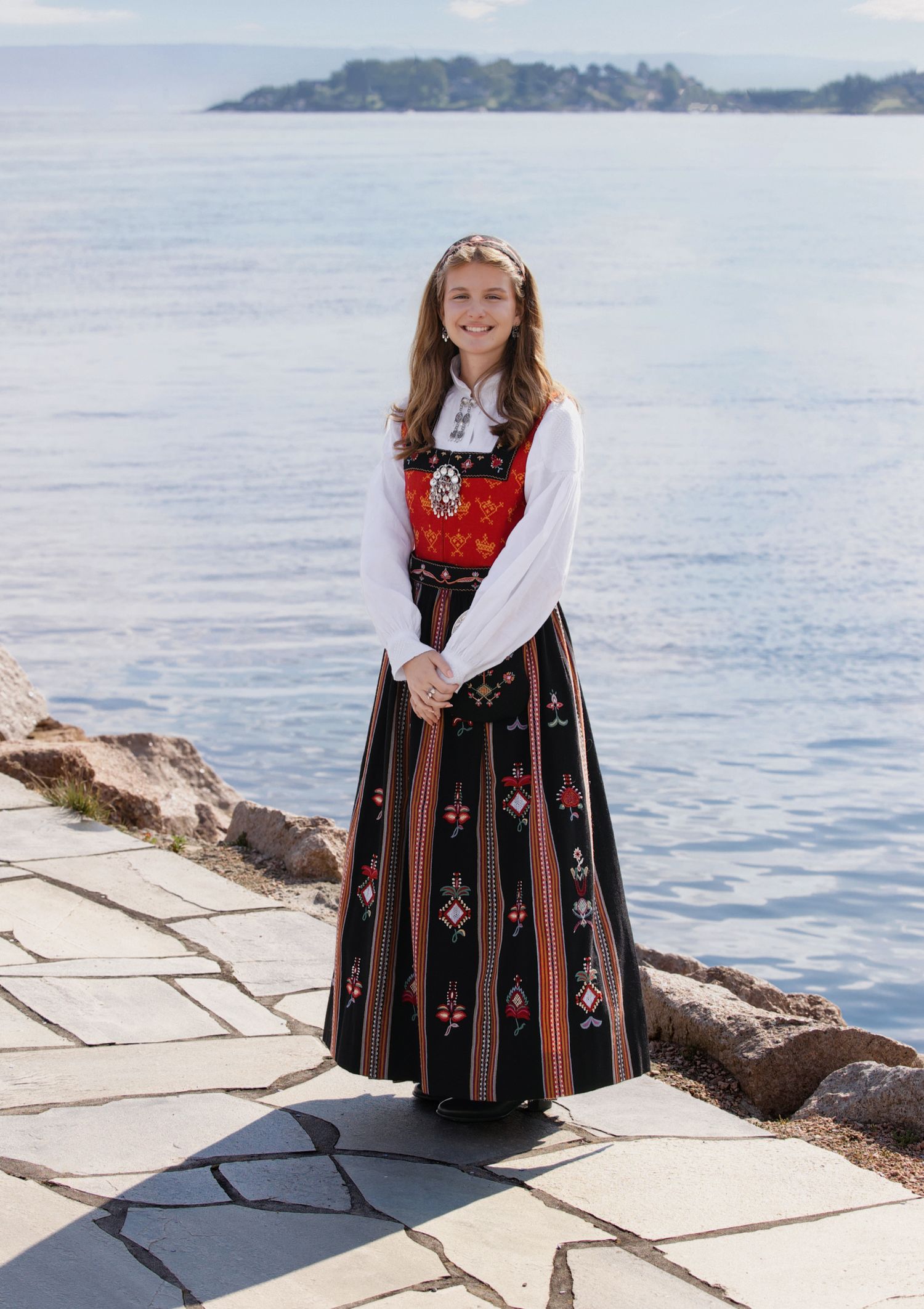 Traditional Norwegian folk costume worn by someone posing by a scenic coastal waterfront with mountains in background.