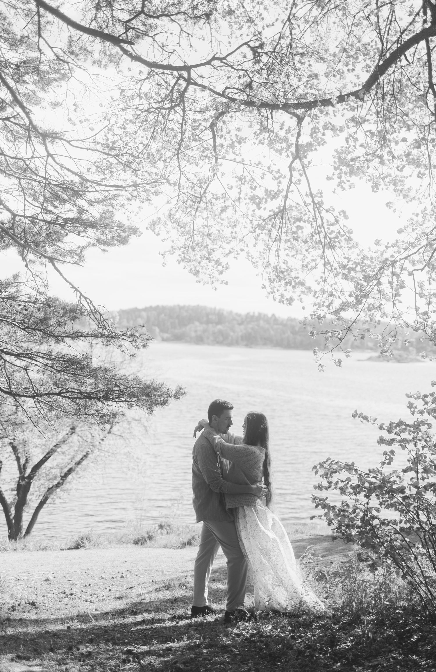 Black and white panoramic photo of couple in wedding attire standing by a serene lake surrounded by trees.