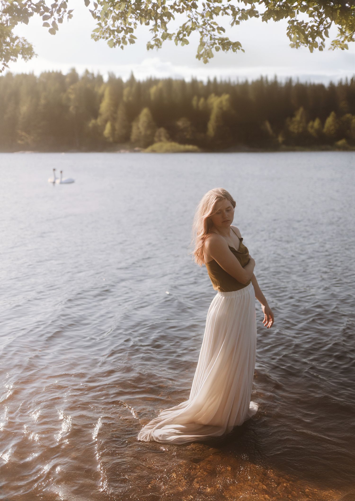 A figure in a flowing white dress stands in shallow lake water at sunset with trees and mountains in the background.