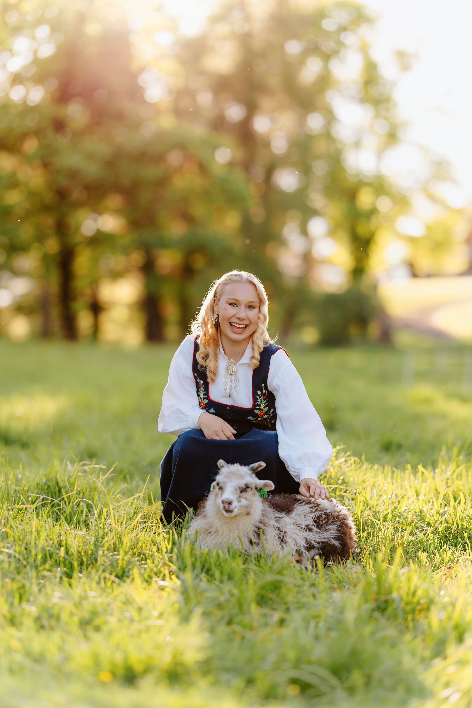 Woman in traditional folk costume smiling while petting a small lamb on a sunny green meadow.