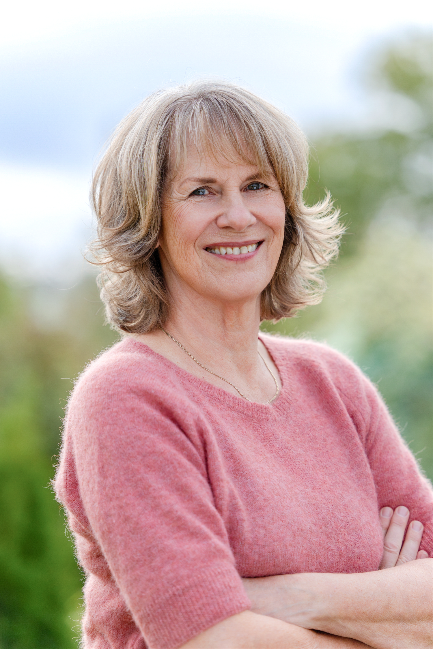 A person wearing a pink sweater poses outdoors with a warm smile against a soft green background.