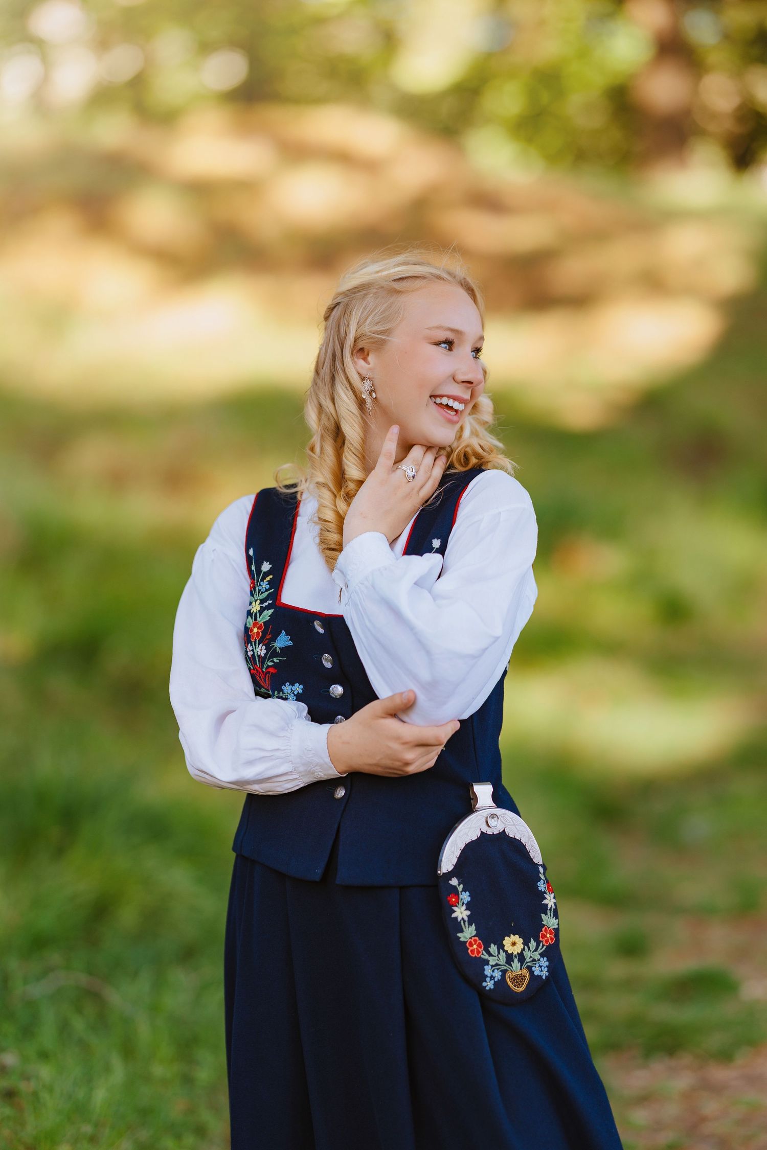 Smiling blonde woman in traditional Norwegian bunad folk costume with floral embroidery, posing outdoors in nature.