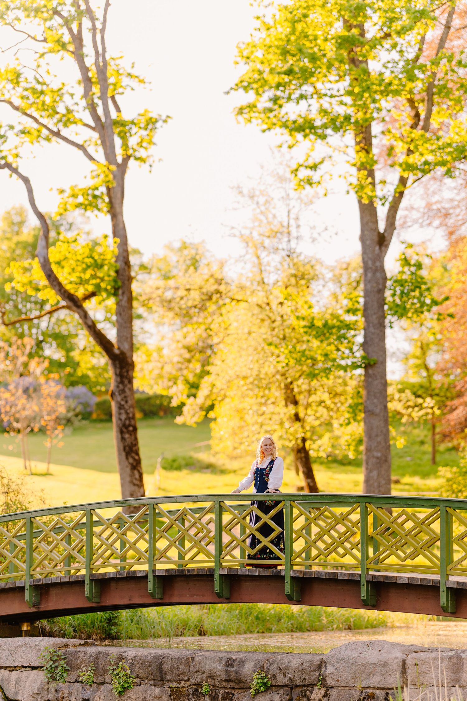 Woman standing on a green ornamental bridge in an autumn park with vibrant yellow foliage.