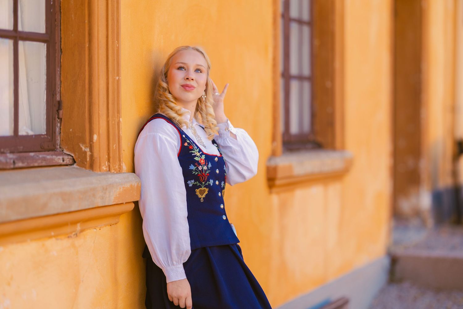 Blonde woman in traditional Norwegian bunad folk costume posing against a yellow building wall outdoors.