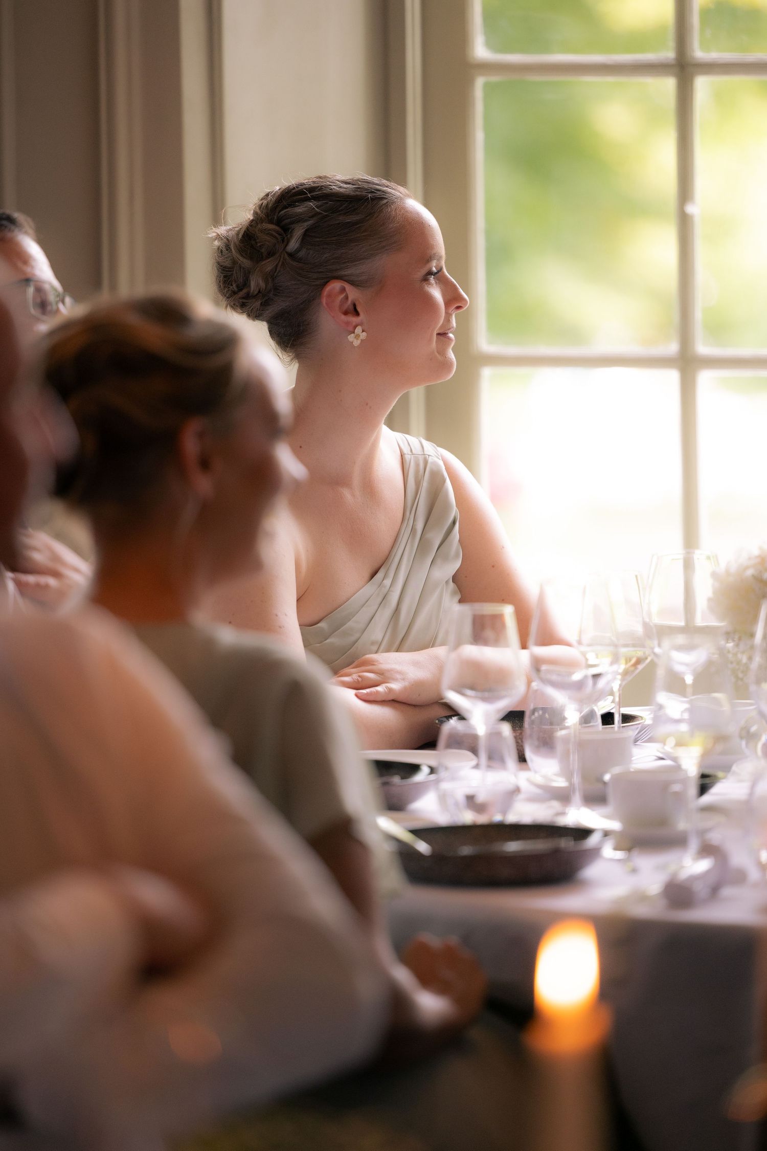 Soft focus candlelit dinner scene showing guests seated at an elegant table in a sunlit dining room.