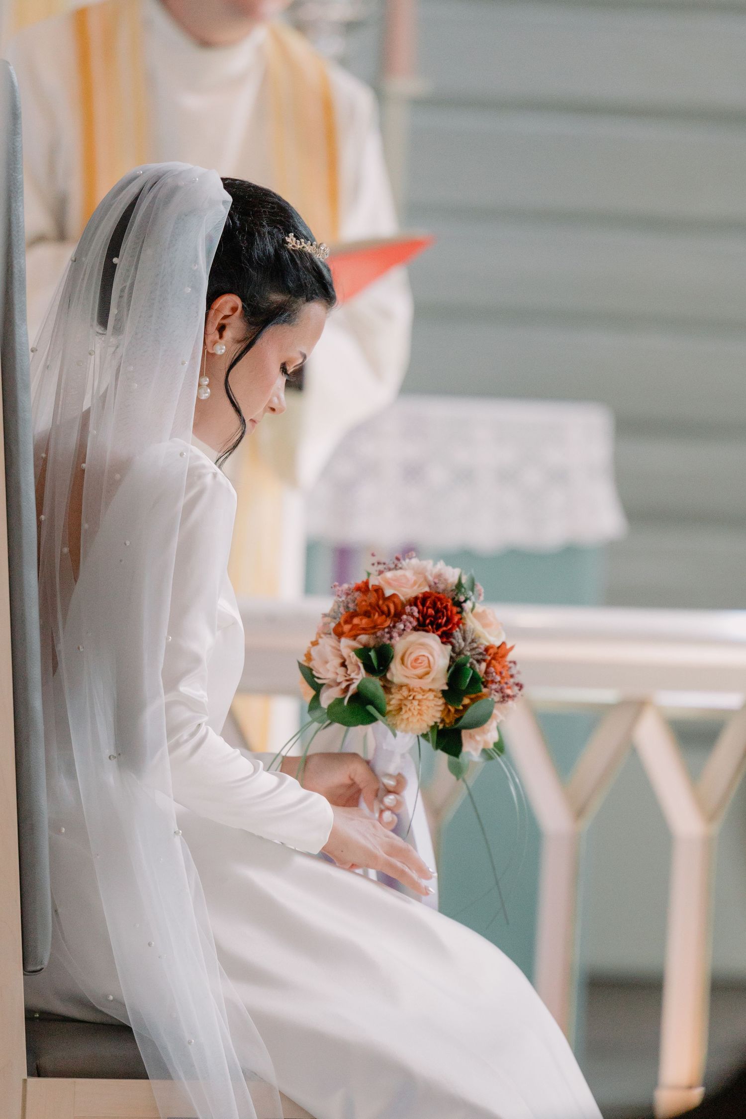 Bride in white wedding dress and veil sits alone with a colorful floral bouquet in a bright interior setting.