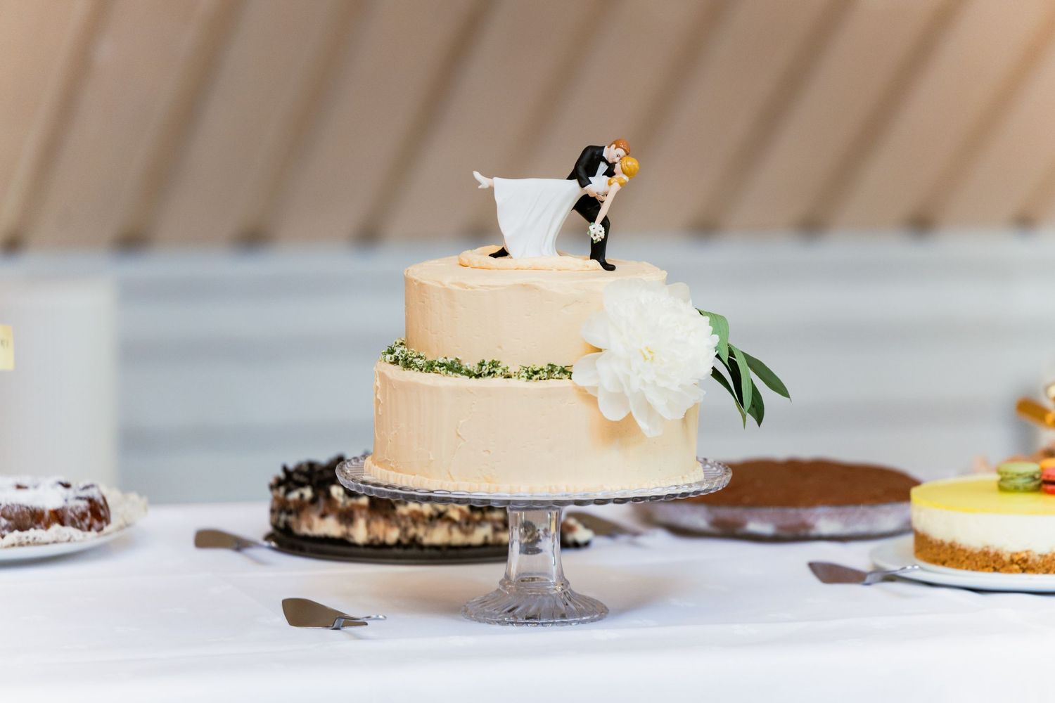 A simple wedding cake decorated with white flowers and a miniature bride and groom topper sits on a silver cake stand.