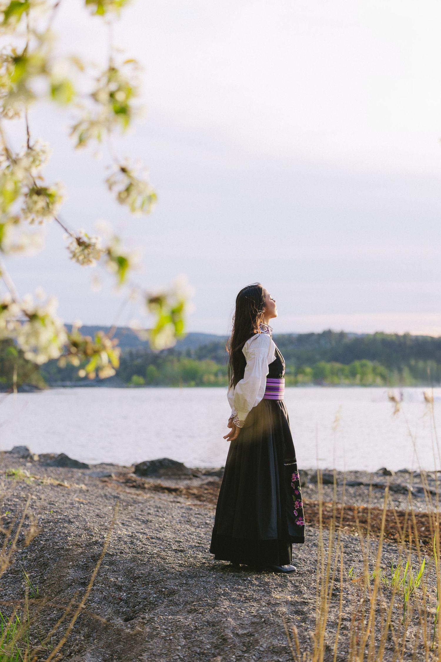 A silhouetted figure in traditional dress walks along a lakeside path with blooming branches in the foreground.