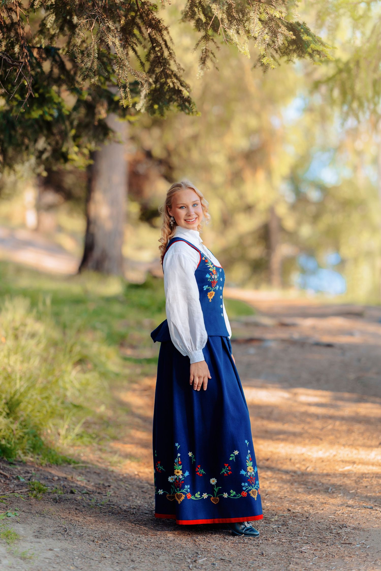 Young woman in traditional Norwegian bunad dress standing on a sunlit forest path surrounded by trees.