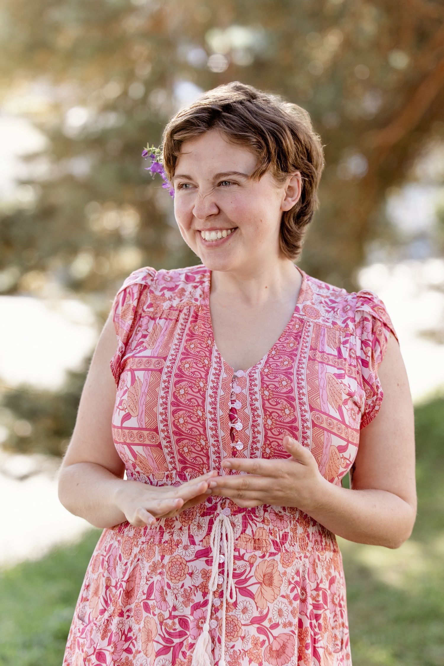 A person in a pink floral summer dress smiles while standing outdoors on a sunny day.