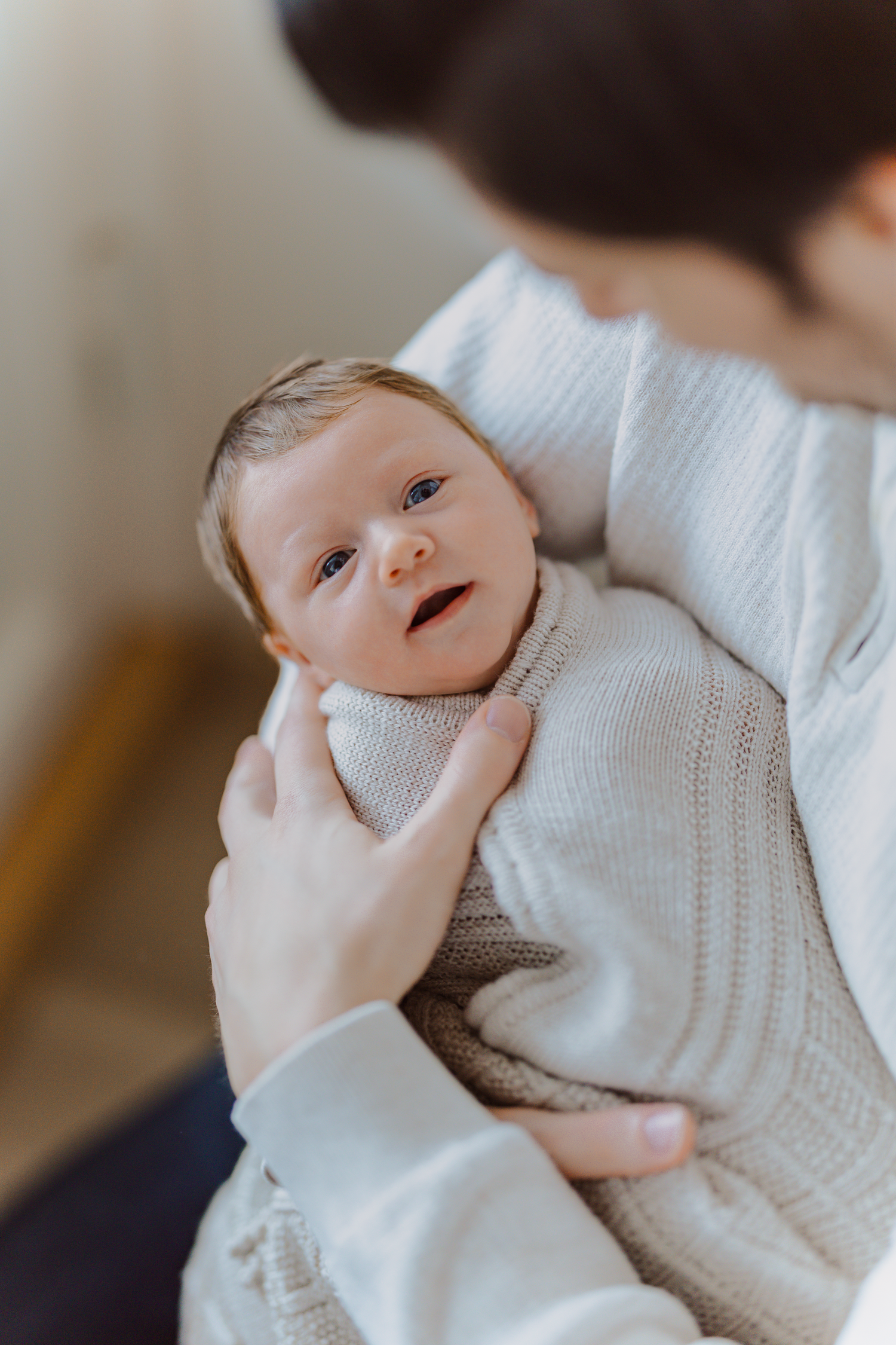 A baby wearing a white knit sweater smiles and laughs while being held.