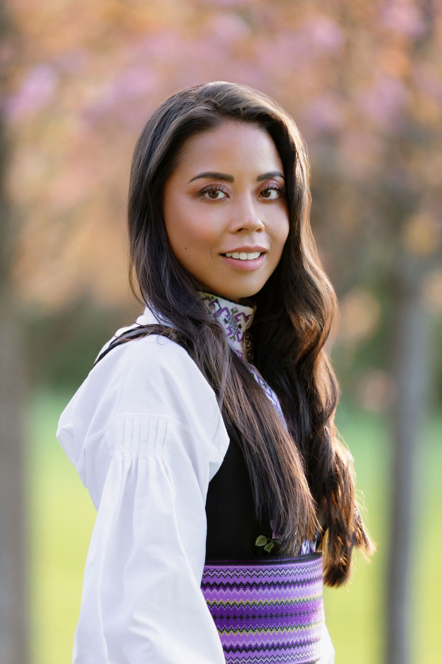 Portrait of a smiling person wearing a white blouse and purple patterned vest against a soft autumn background.