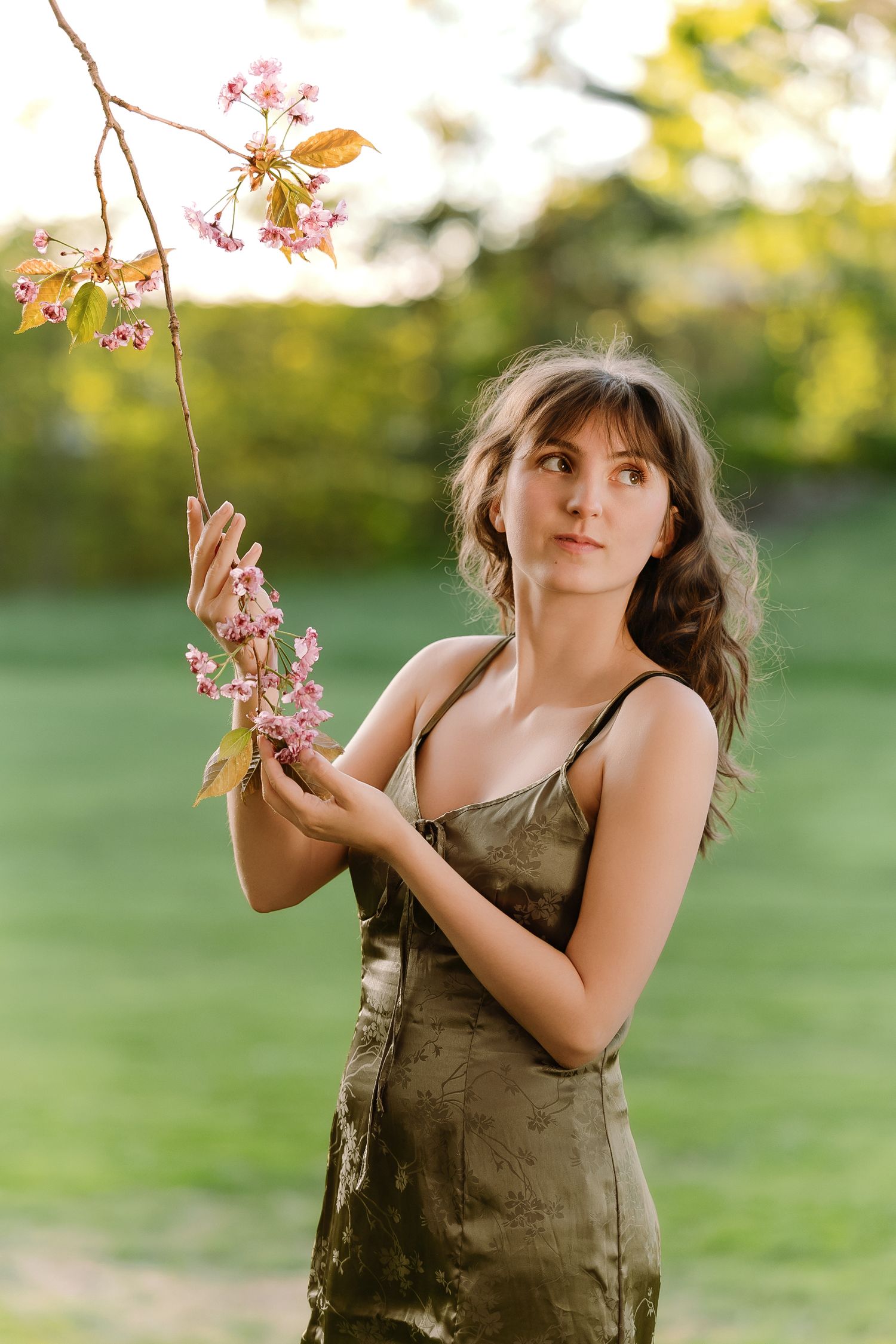 A person in an olive dress poses outdoors near delicate pink cherry blossoms on a spring day.