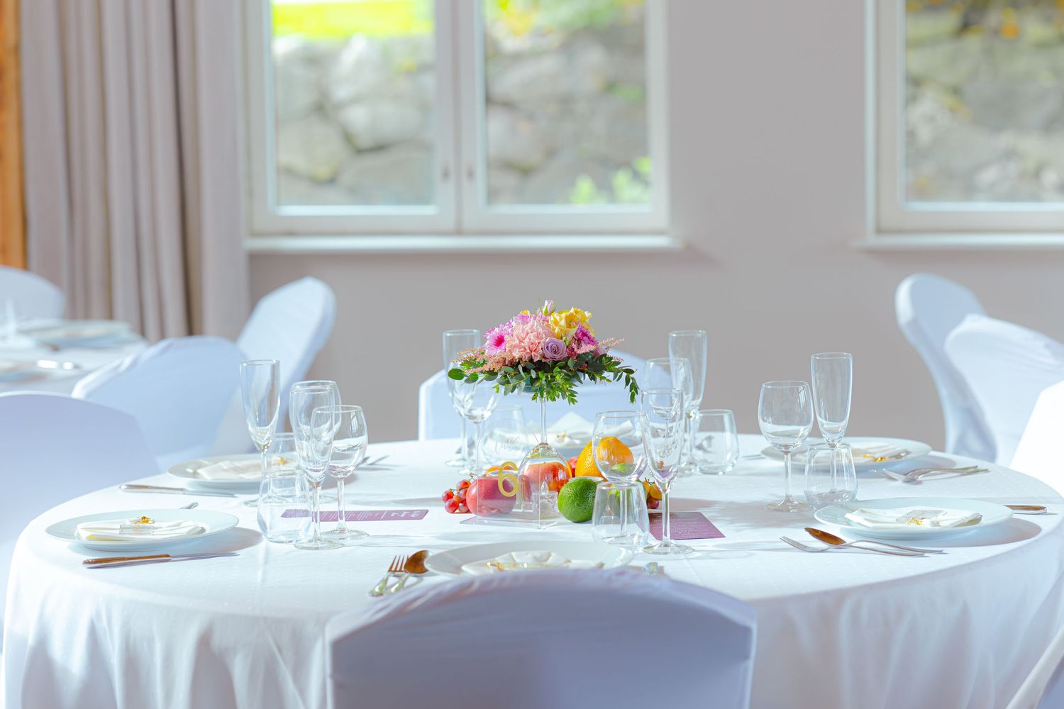 Elegant wedding reception table setting with white linens, wine glasses, and a tiered floral centerpiece in natural light.