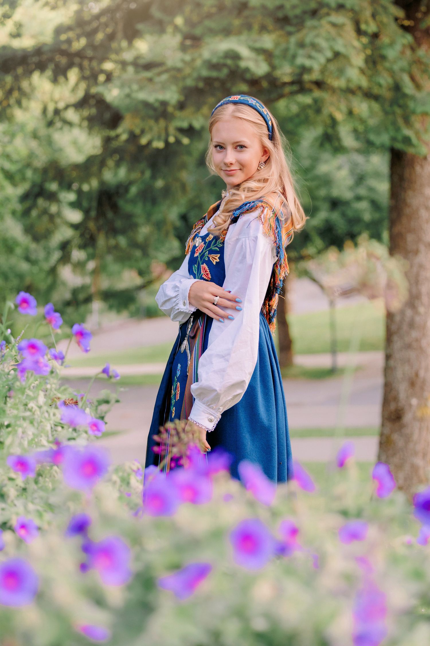 A person in traditional Scandinavian folk dress poses in a garden setting with vibrant purple wildflowers in the foreground.