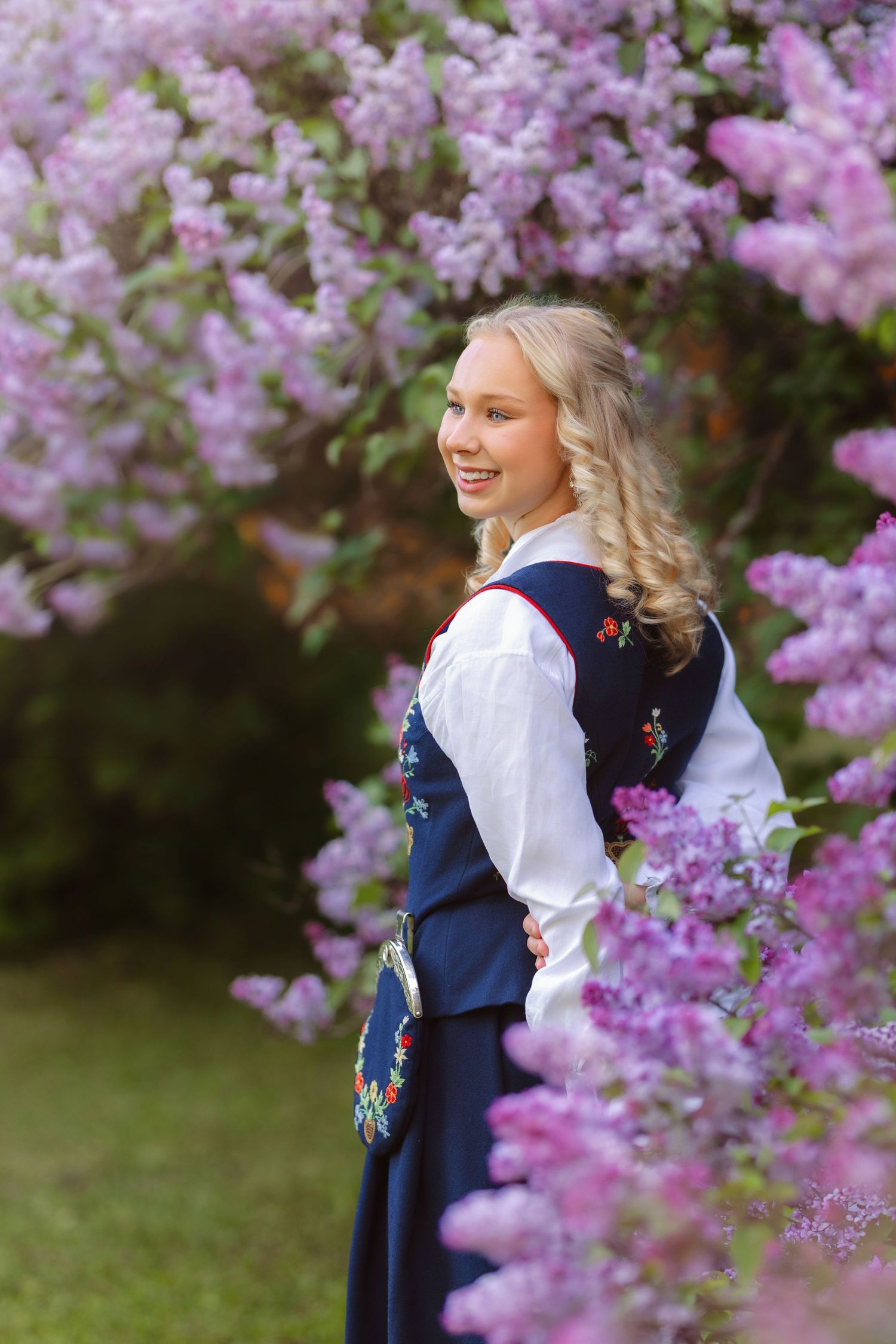 Young blonde girl in traditional Norwegian bunad dress smiling among blooming lilac flowers outdoors.