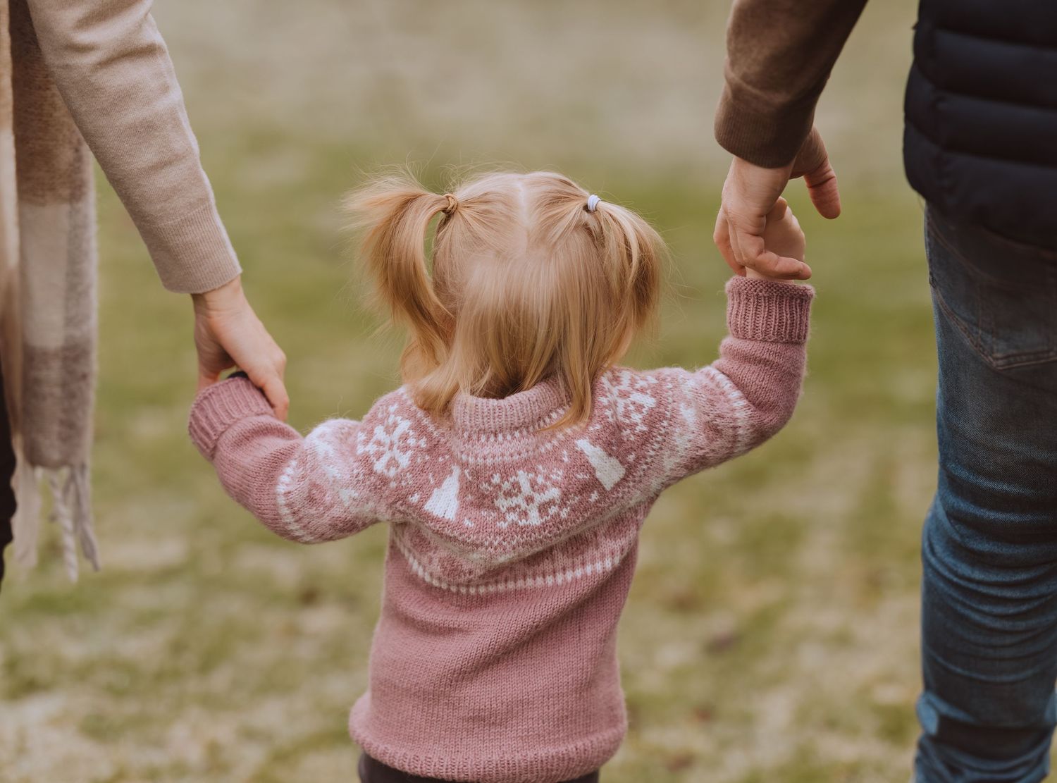 A family holds hands while walking together outdoors, wearing cozy sweaters in pink and neutral tones.