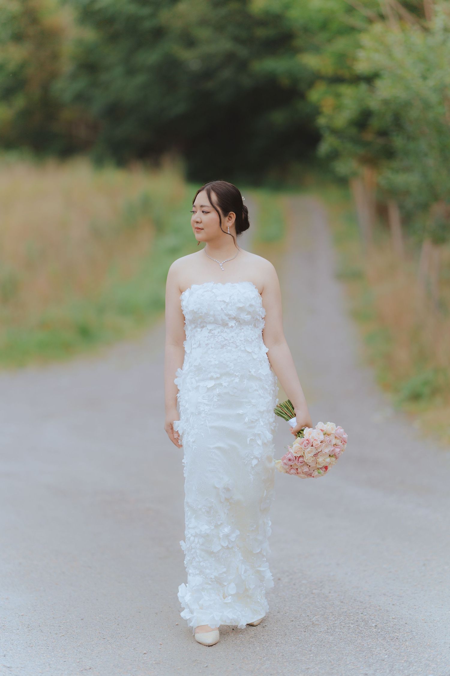 A bride in a white textured dress walks down a gravel path holding a pink bouquet in a romantic outdoor setting.