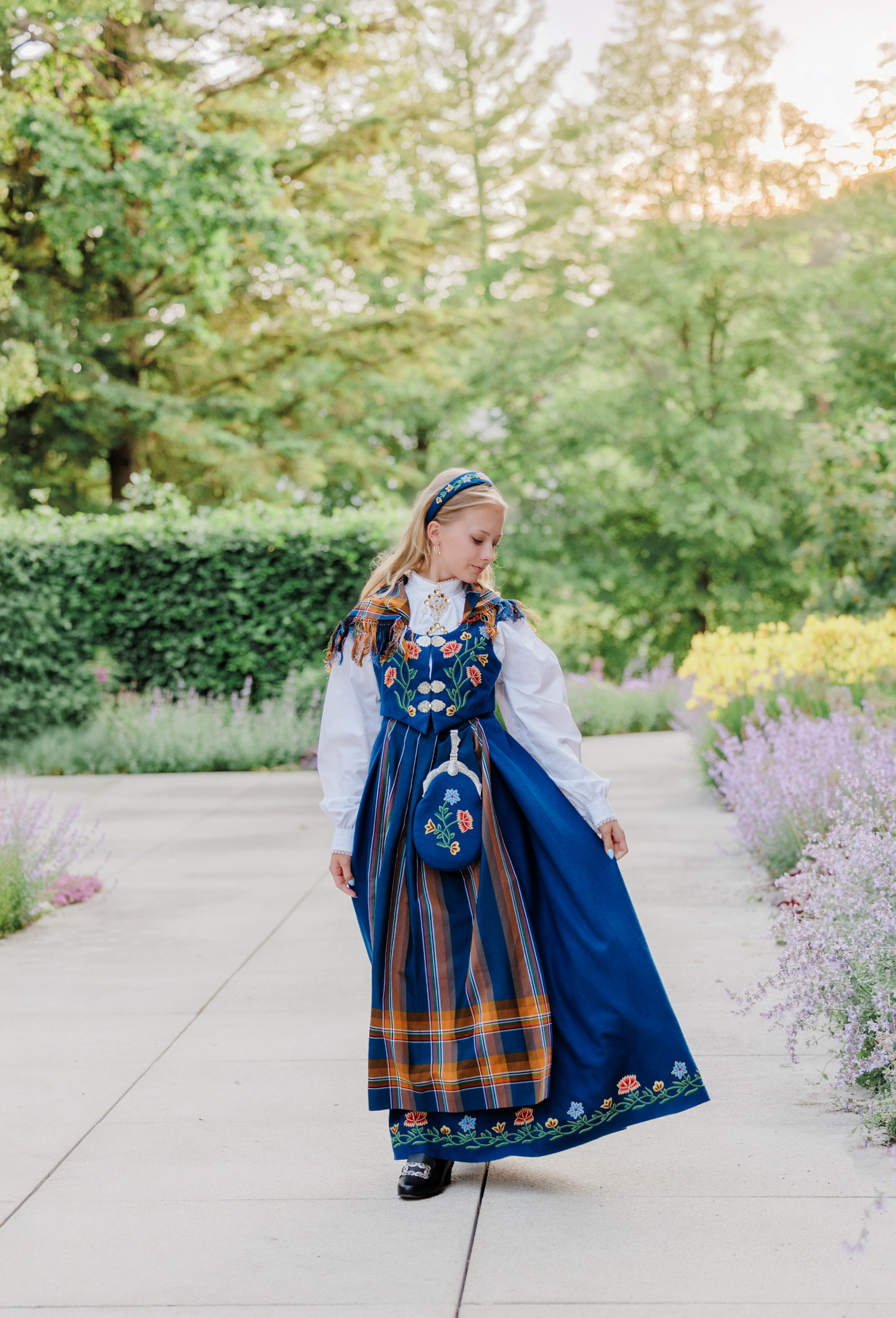 Traditional Norwegian folk costume in navy blue and orange with embroidered details shown in outdoor garden setting.