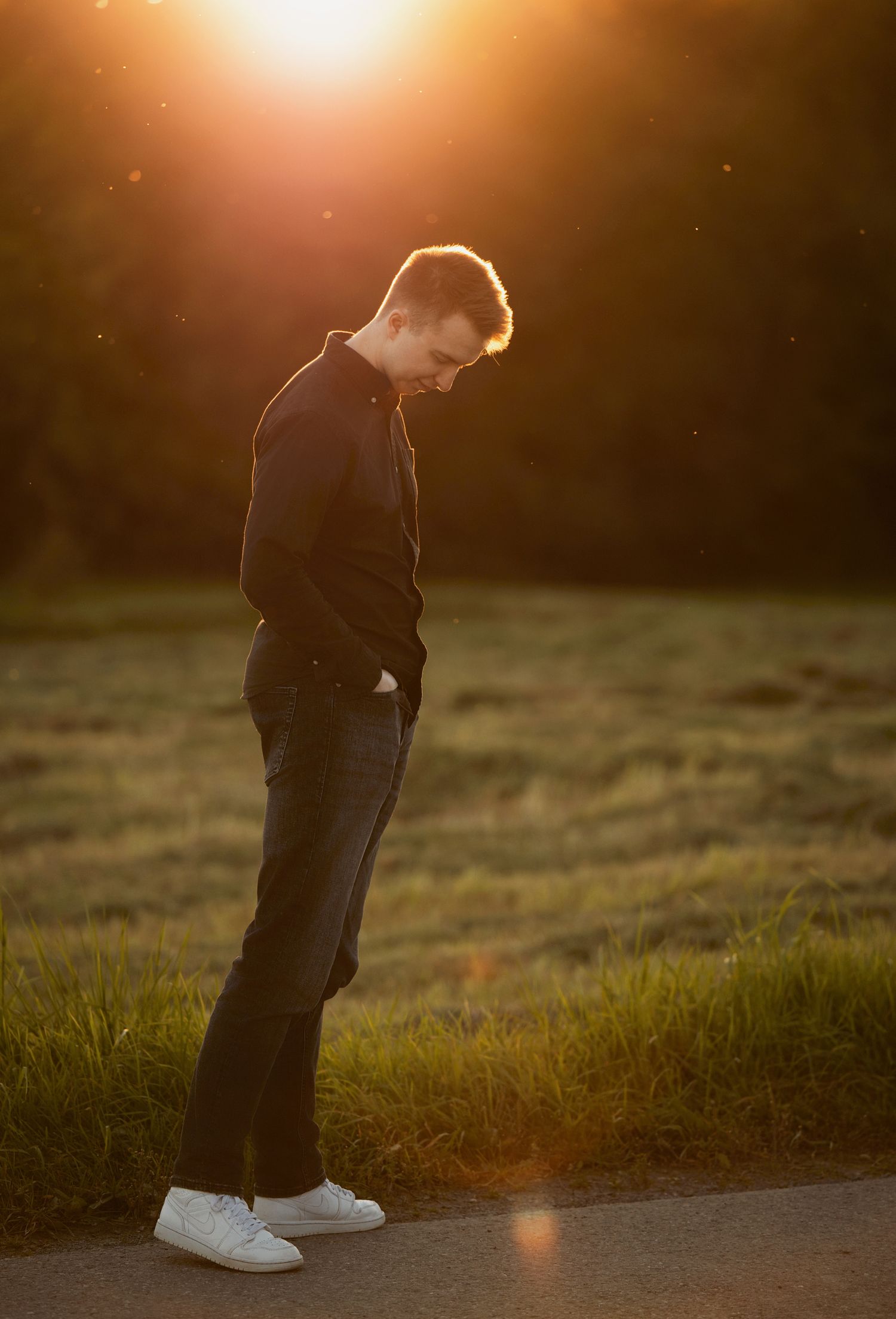 A small figure stands silhouetted against a golden sunset on a grassy hillside wearing a dark jacket and white shoes.