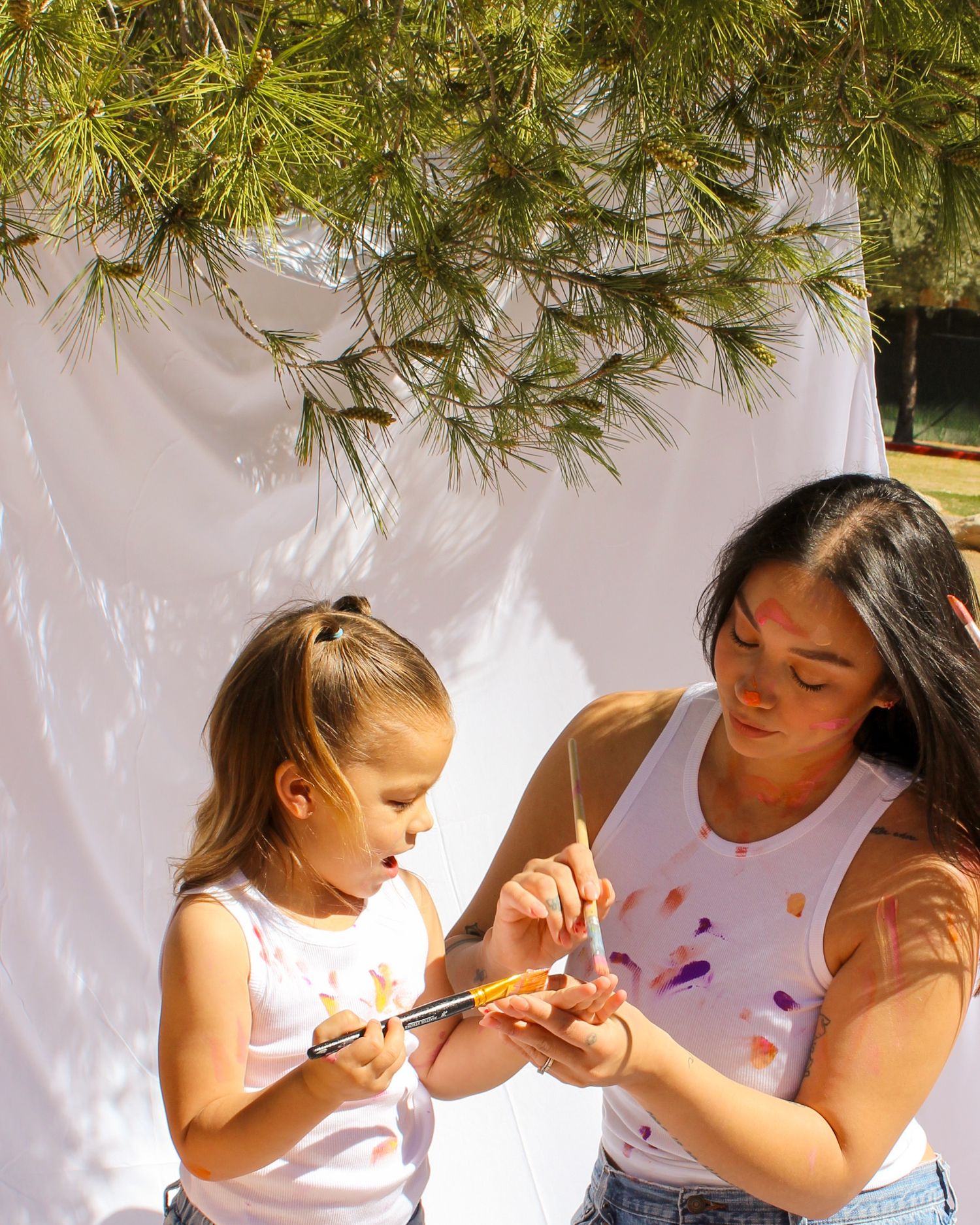 Mother and daughter painting together outdoors near white backdrop under pine tree in sunlight.