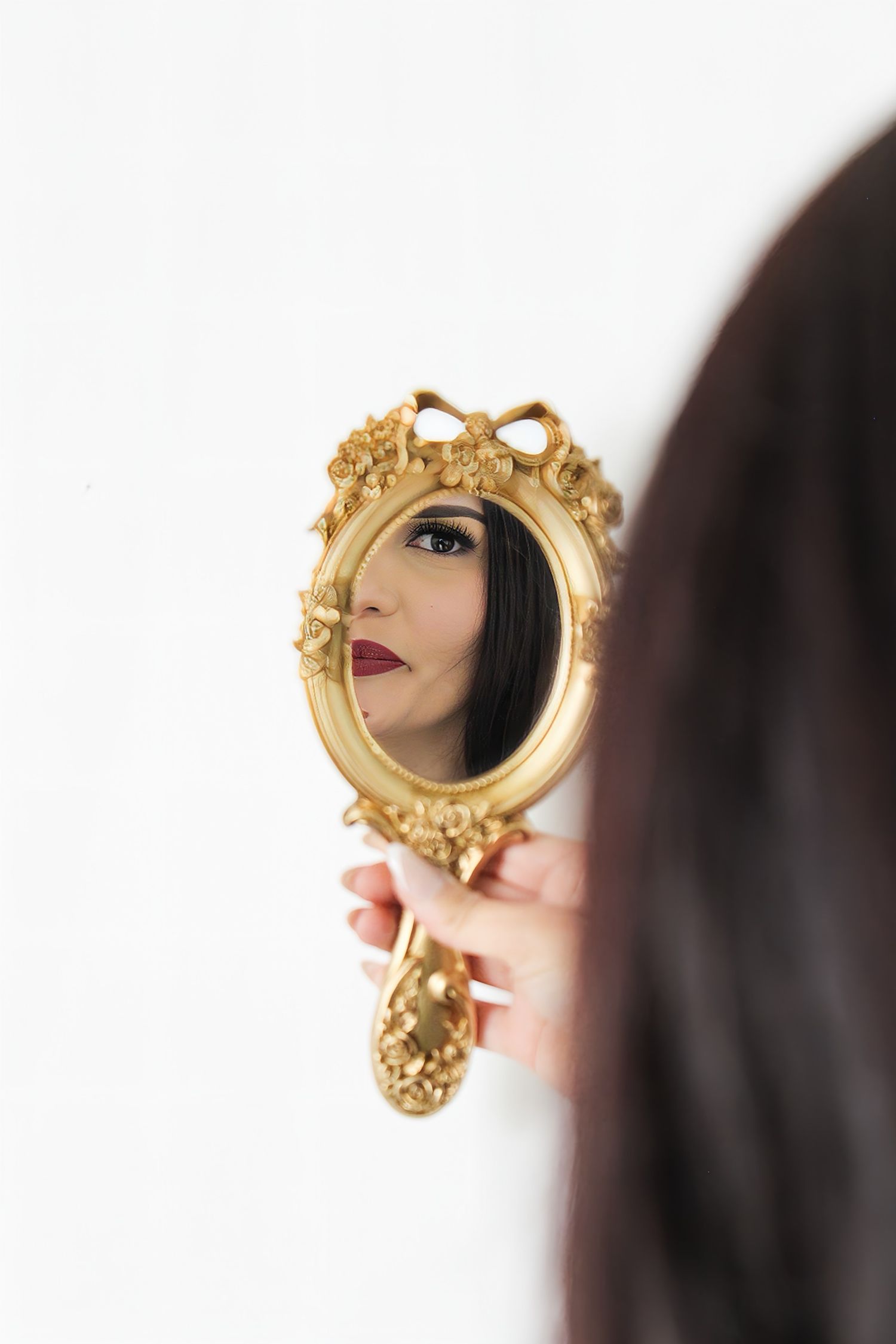 Woman holding an ornate gold oval hand mirror reflecting her face with red lips against a white wall.