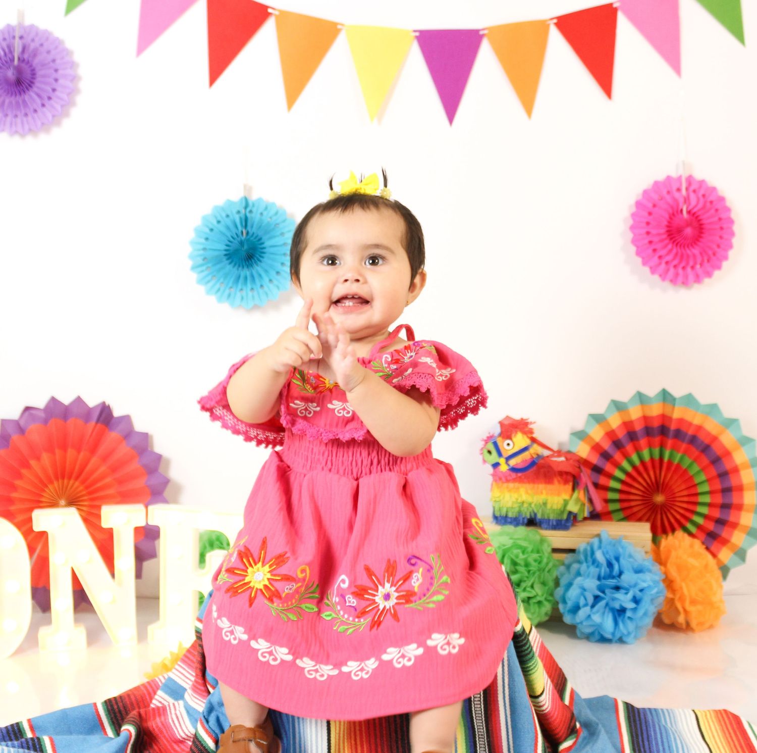 Baby girl in pink floral dress at colorful fiesta first birthday party with pinata and paper fans.